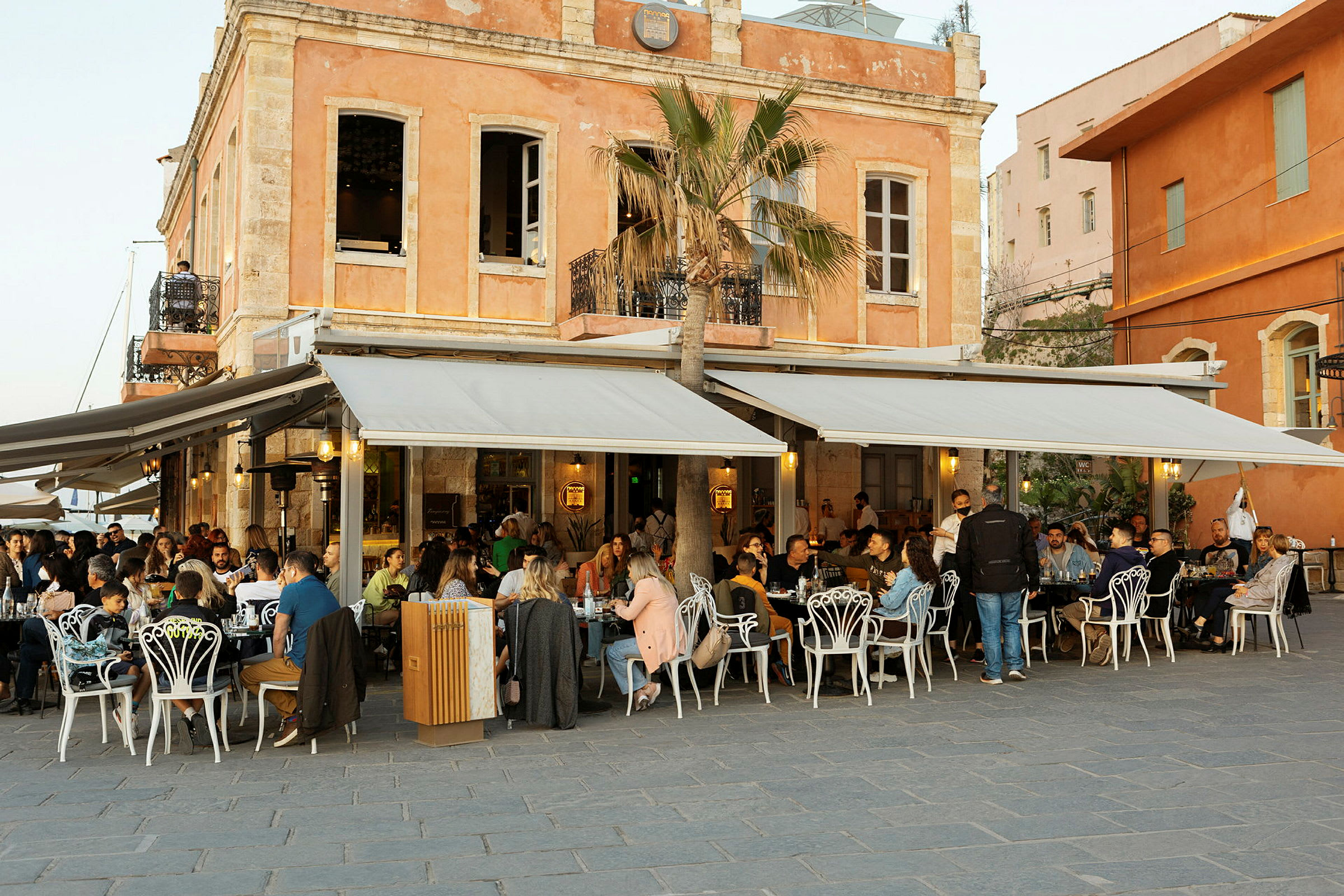 Folk sidder og spiser på en café med udendørs servering foran en bygning med orange facade. (AI-genereret tekst)