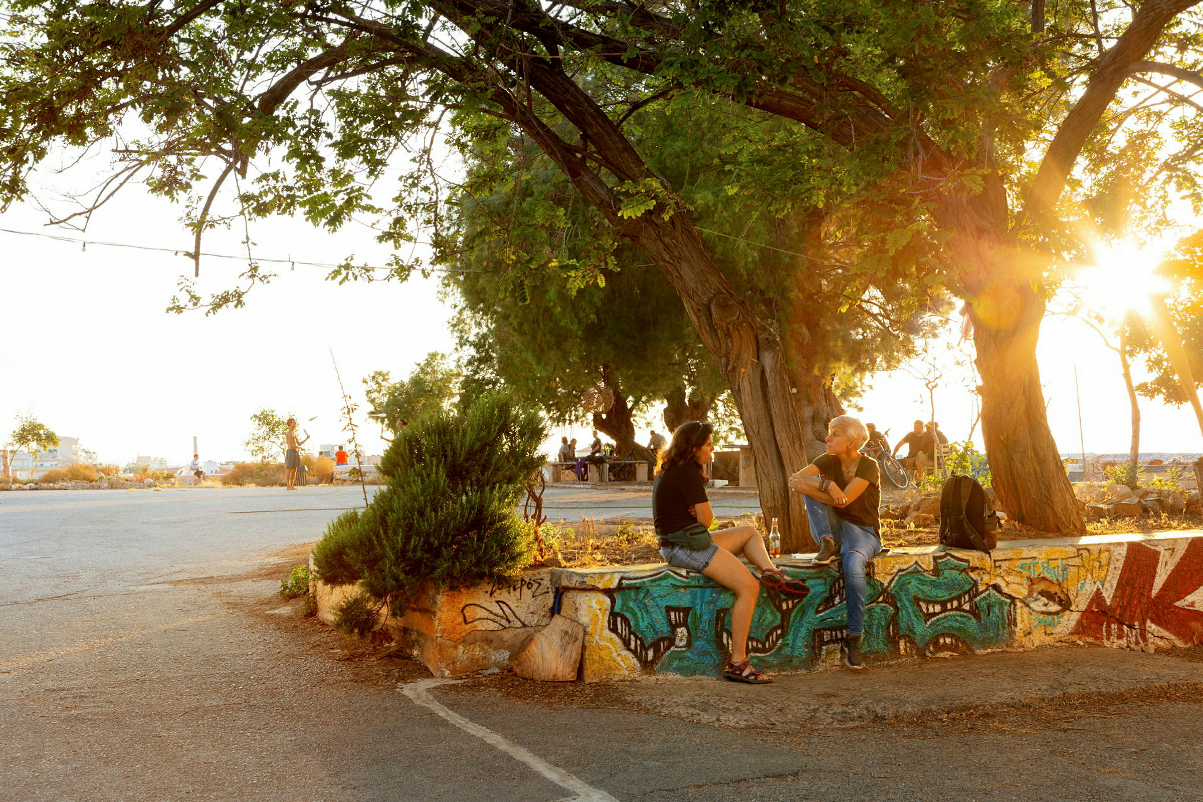 Personer sidder på en graffitimalet mur under et træ, med solnedgang i baggrunden. (AI-genereret tekst)
