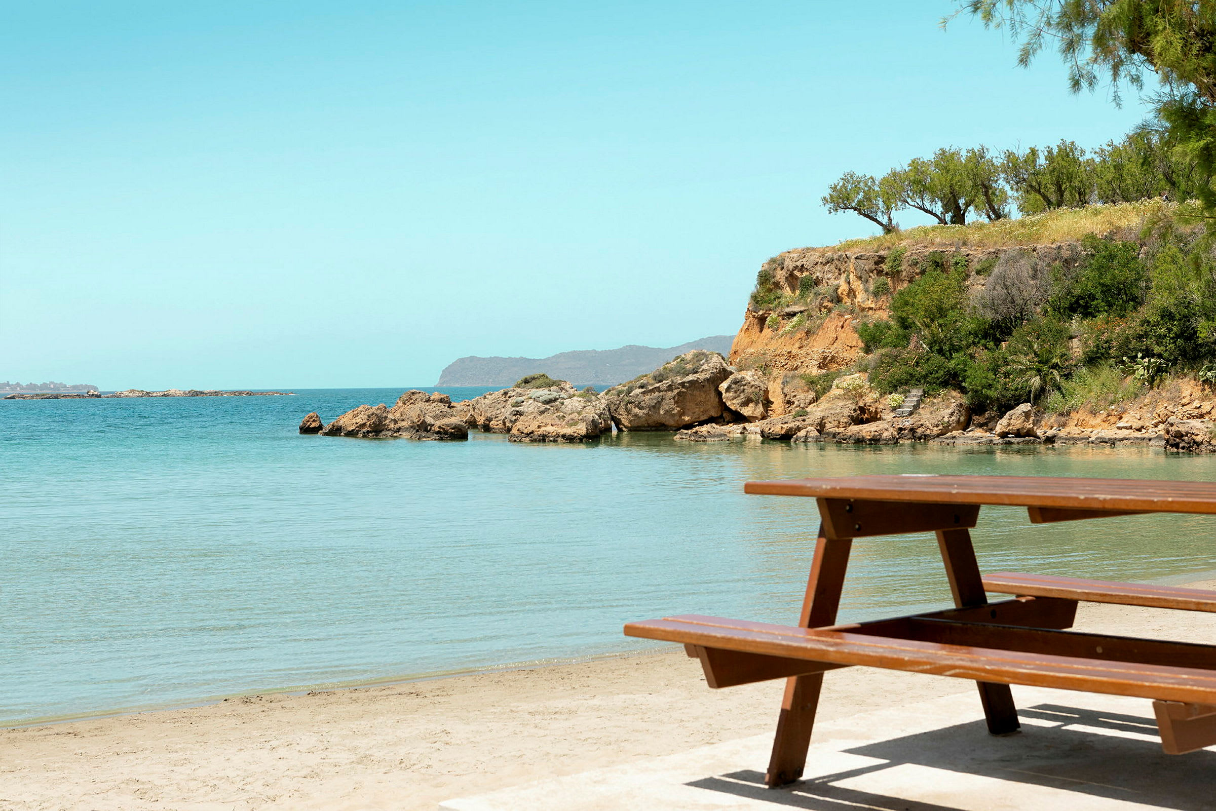 Picnicbord ved en sandstrand med klart vand og klippeformationer i baggrunden. (AI-genereret tekst)