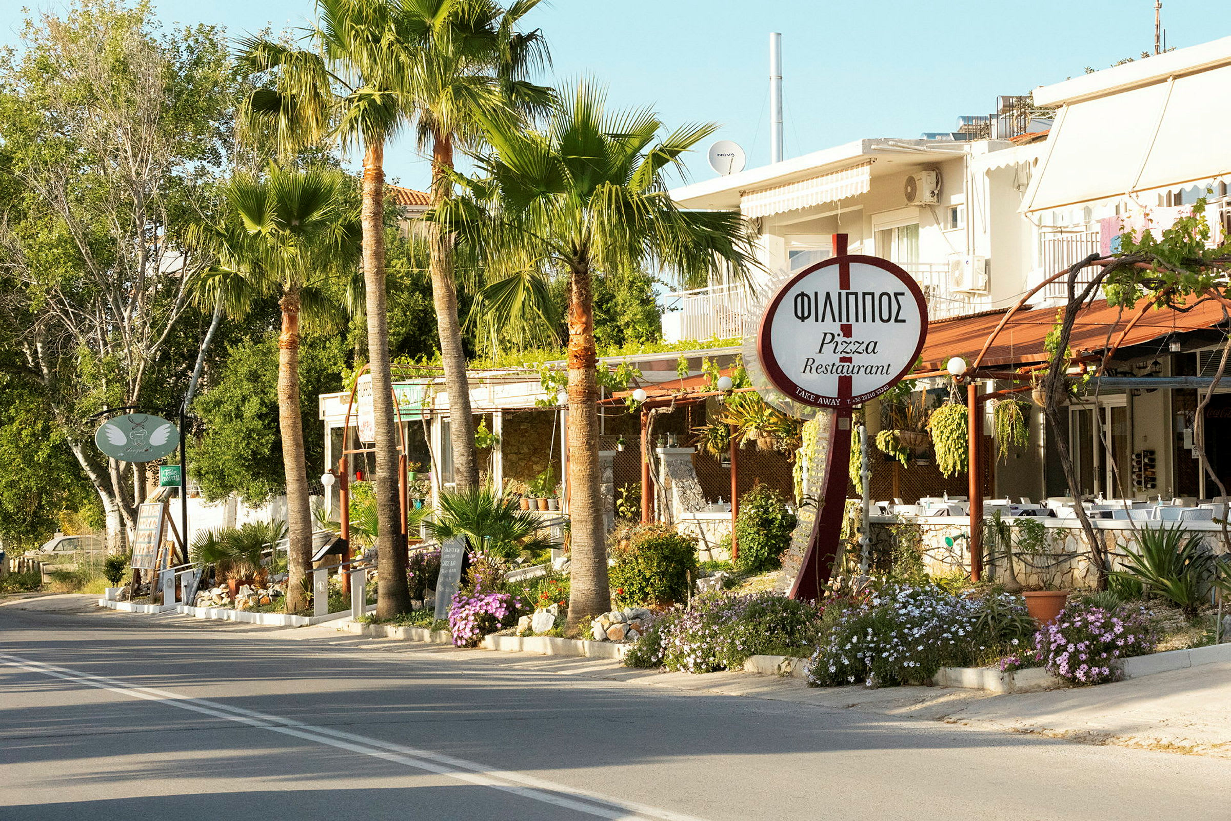 En vej kantet med palmer passerer forbi en restaurant med et skilt, der viser navnet 'Philippos Pizza Restaurant'. (AI-genereret tekst)