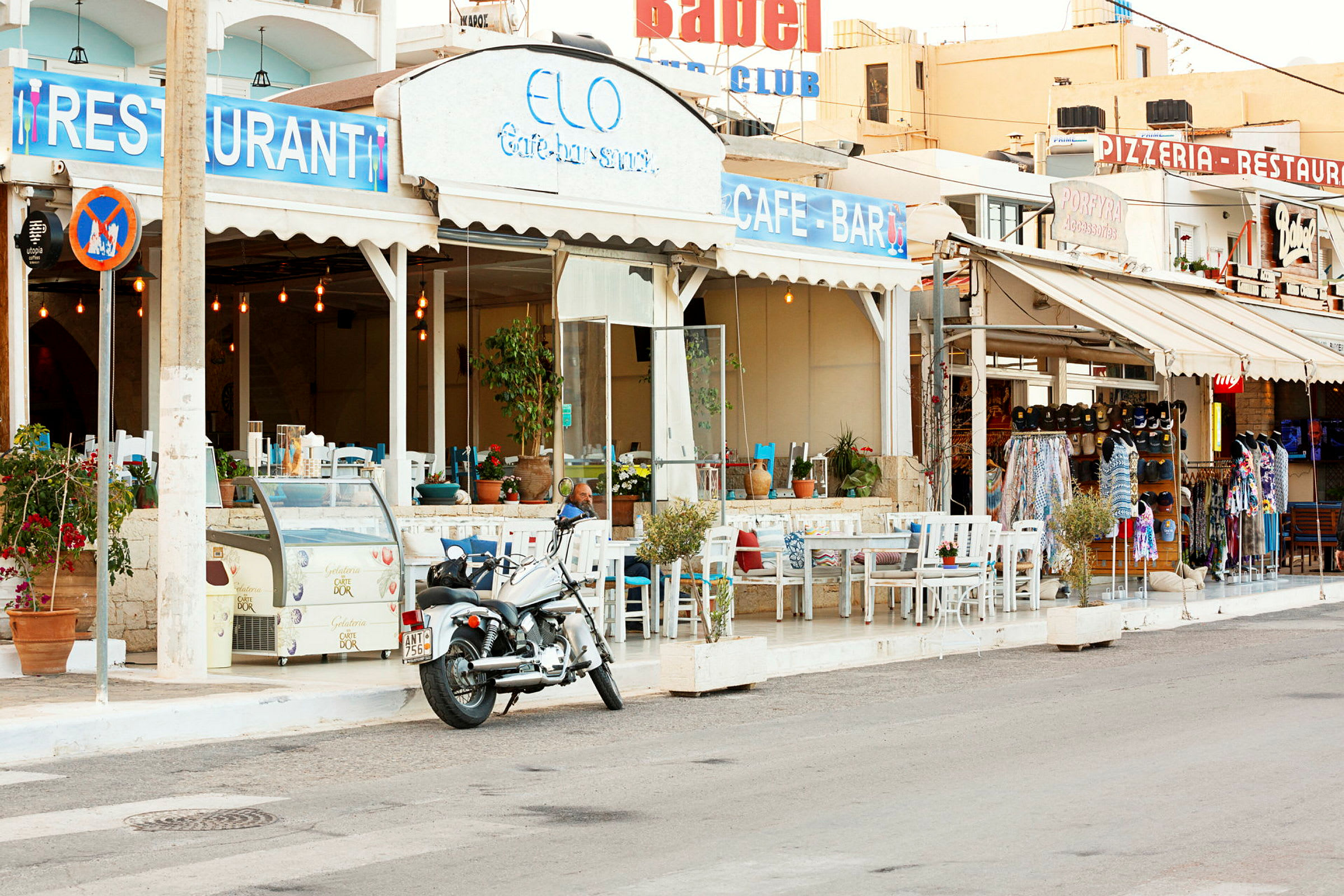 Motiv af en café med udendørs servering ved en gade, et skilt med teksten "Restaurant" og en motorcykel parkeret i forgrunden. (AI-genereret tekst)