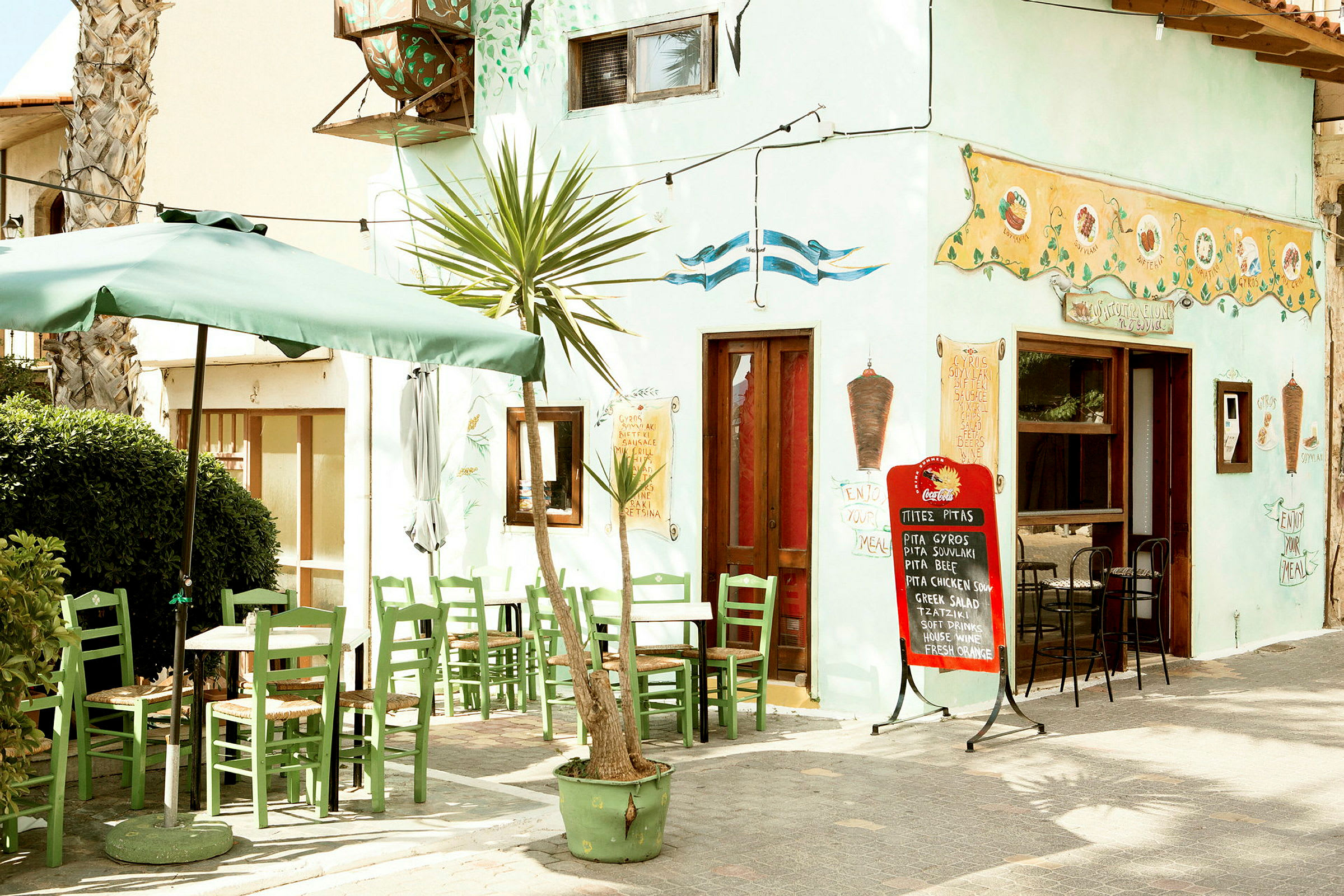 En lille restaurant med grønne stole og borde udendørs under en parasol, med menu skrevet på en skilt. (AI-genereret tekst)
