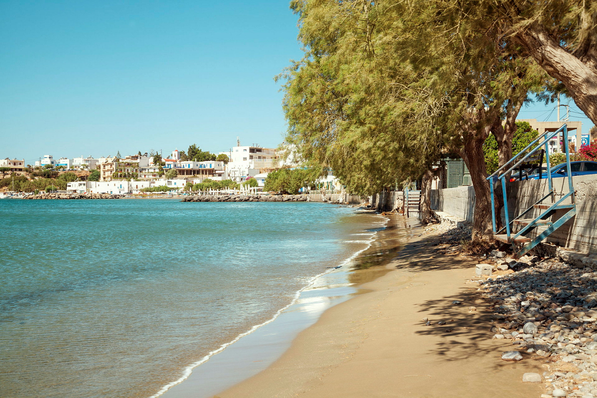 Strand med sand og sten, træer og trin, med en lille by og hav i baggrunden. (AI-genereret tekst)