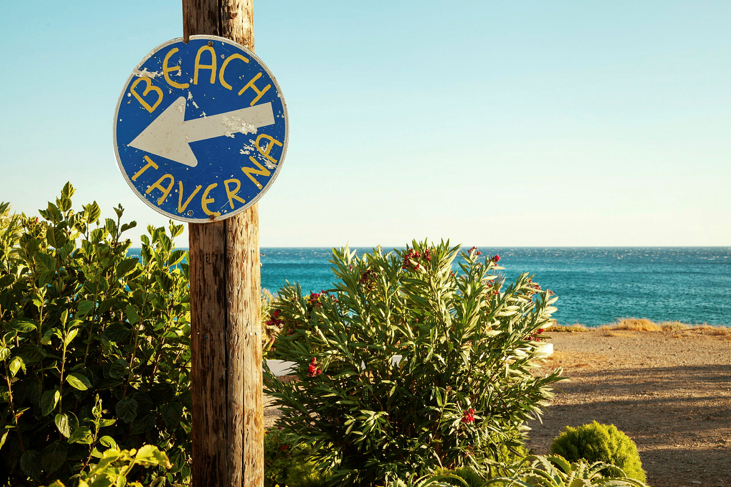 Et skilt med teksten 'Beach Taverna' peger mod stranden, med vegetation og havet i baggrunden. (AI-genereret tekst)