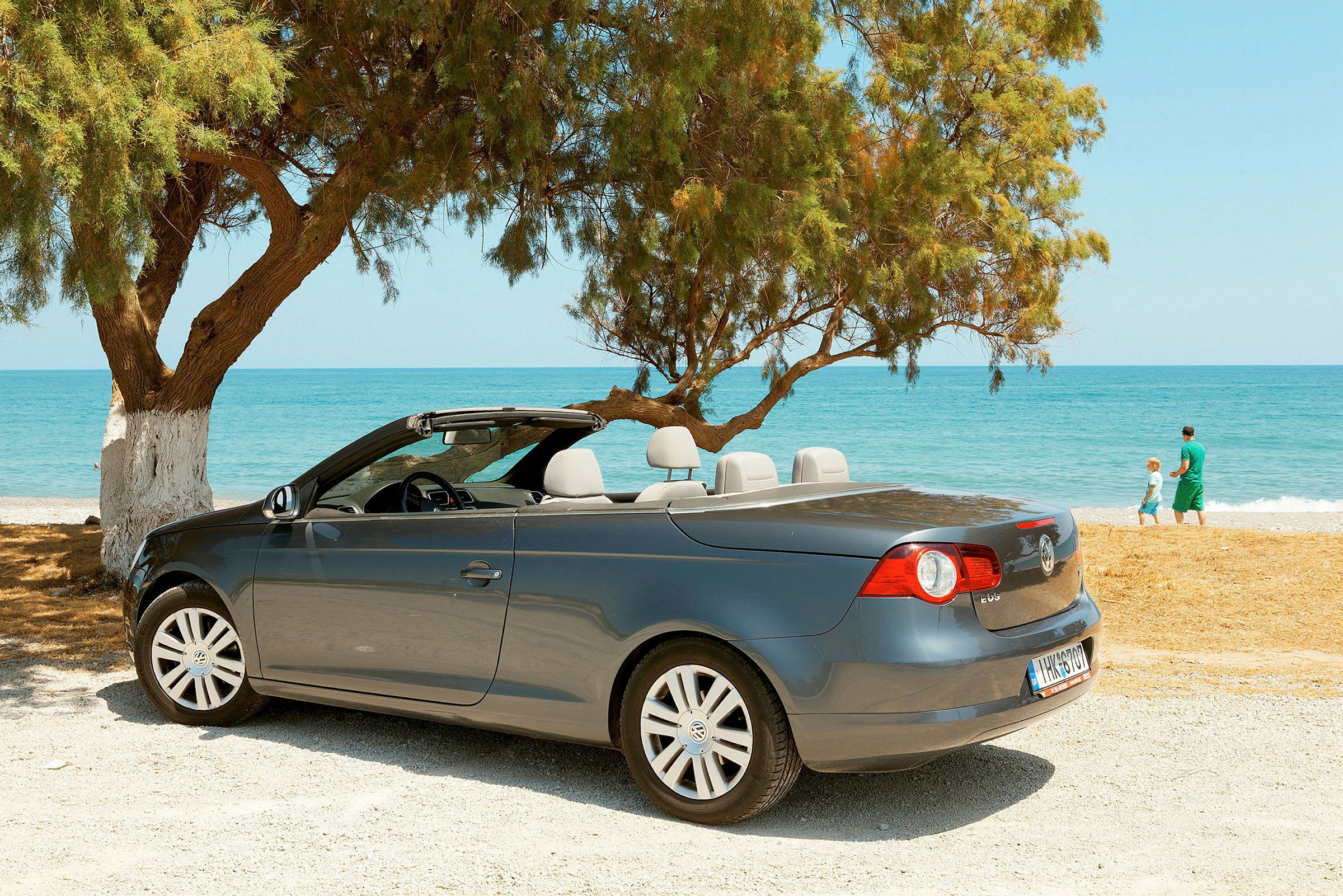 En grå cabriolet er parkeret ved en strand med hav og træer i baggrunden. (AI-genereret tekst)