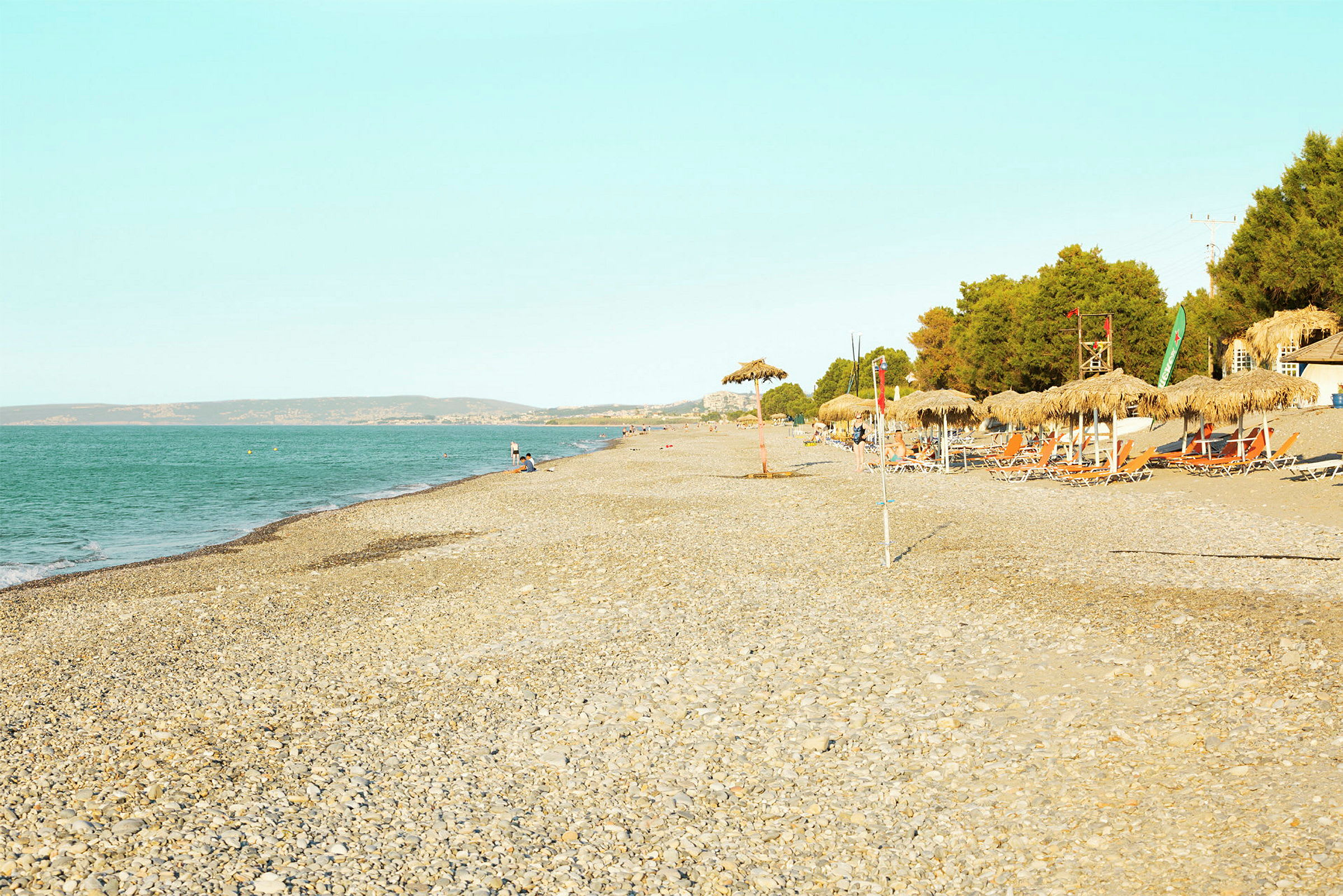 En strand med sten, parasoller og liggestole ved siden af en skov. Havet er synligt til venstre. (AI-genereret tekst)