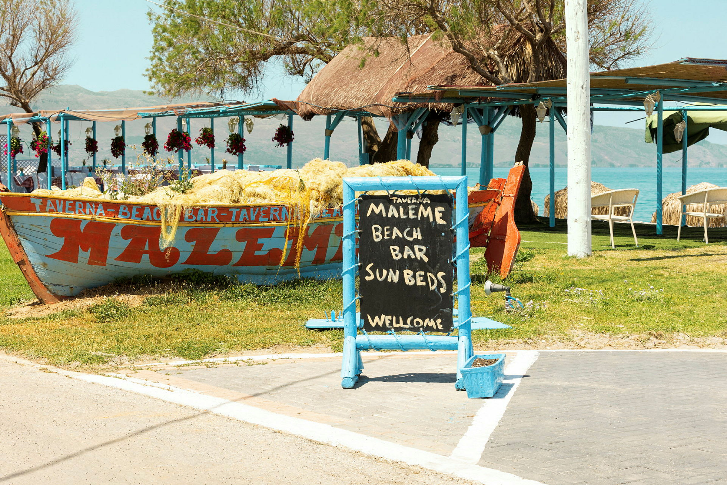 Skilt med teksten 'Maleme Beach Bar Sunbeds Welcome' foran en båd dekoreret med net samt udendørs servering ved havet. (AI-genereret tekst)