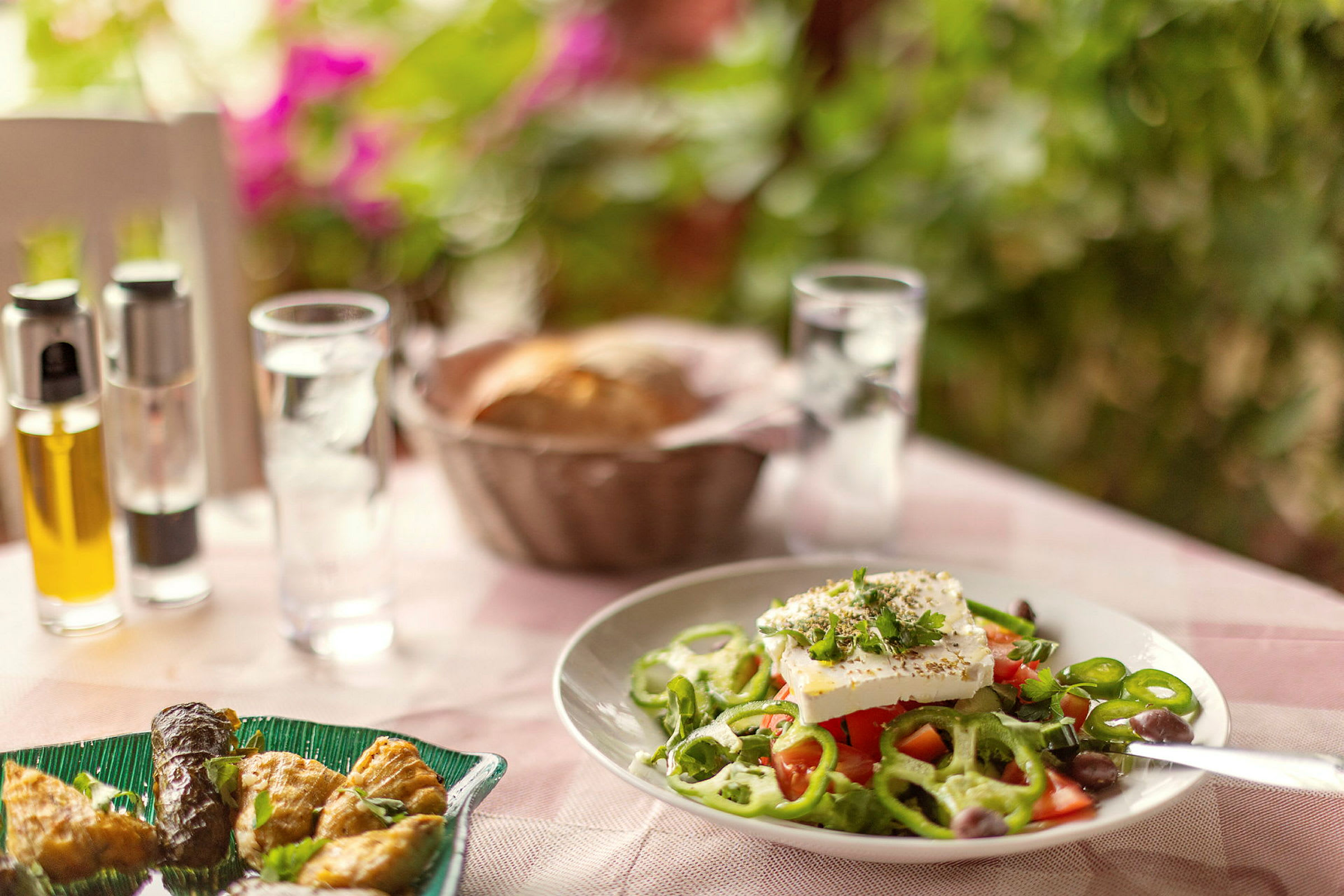 En tallerken med græsk salat på et bord, med olie, peber og brødkurv i baggrunden. (AI-genereret tekst)