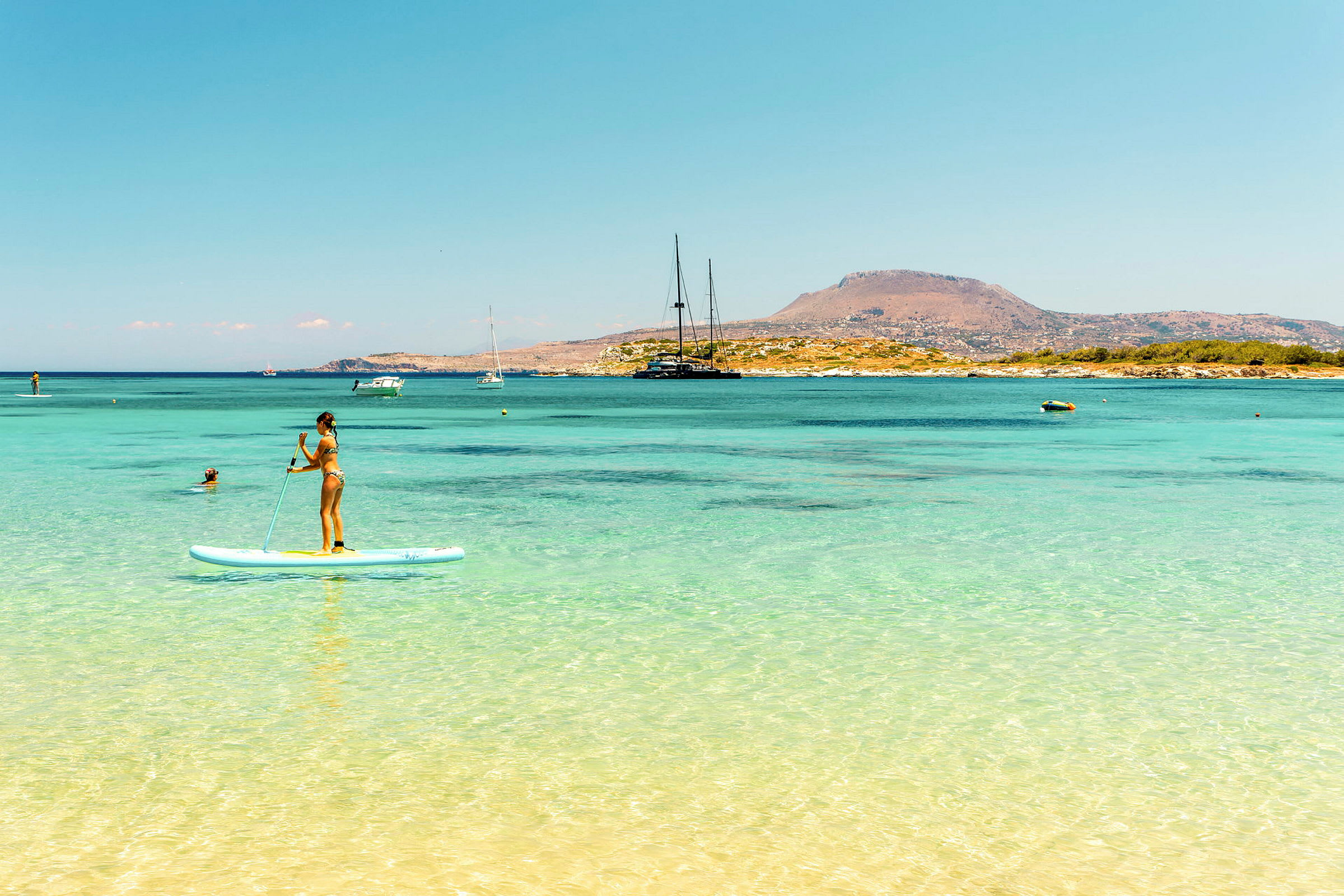 Person, der står på et paddleboard i klart blåt vand med en bjergfyldt kystlinje i baggrunden. (AI-genereret tekst)