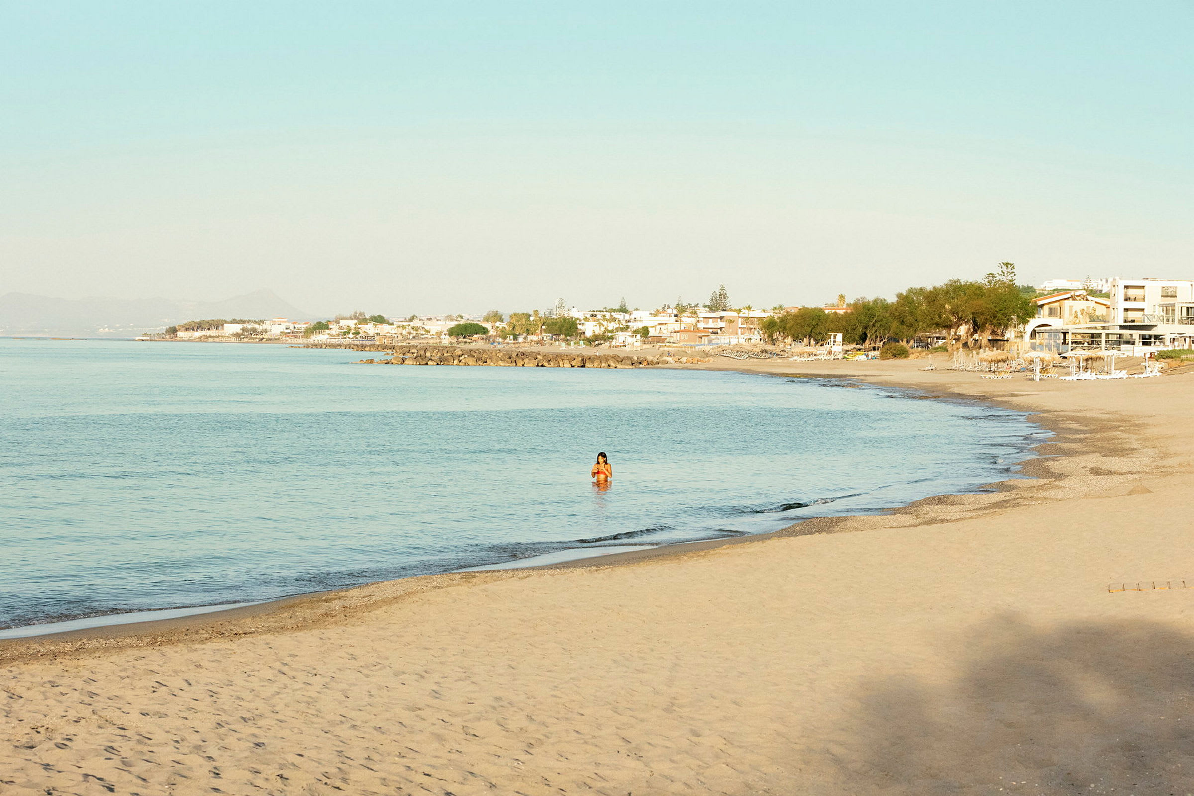 En sandstrand med roligt blåt vand, en person er synlig i vandet, og huse i baggrunden. (AI-genereret tekst)