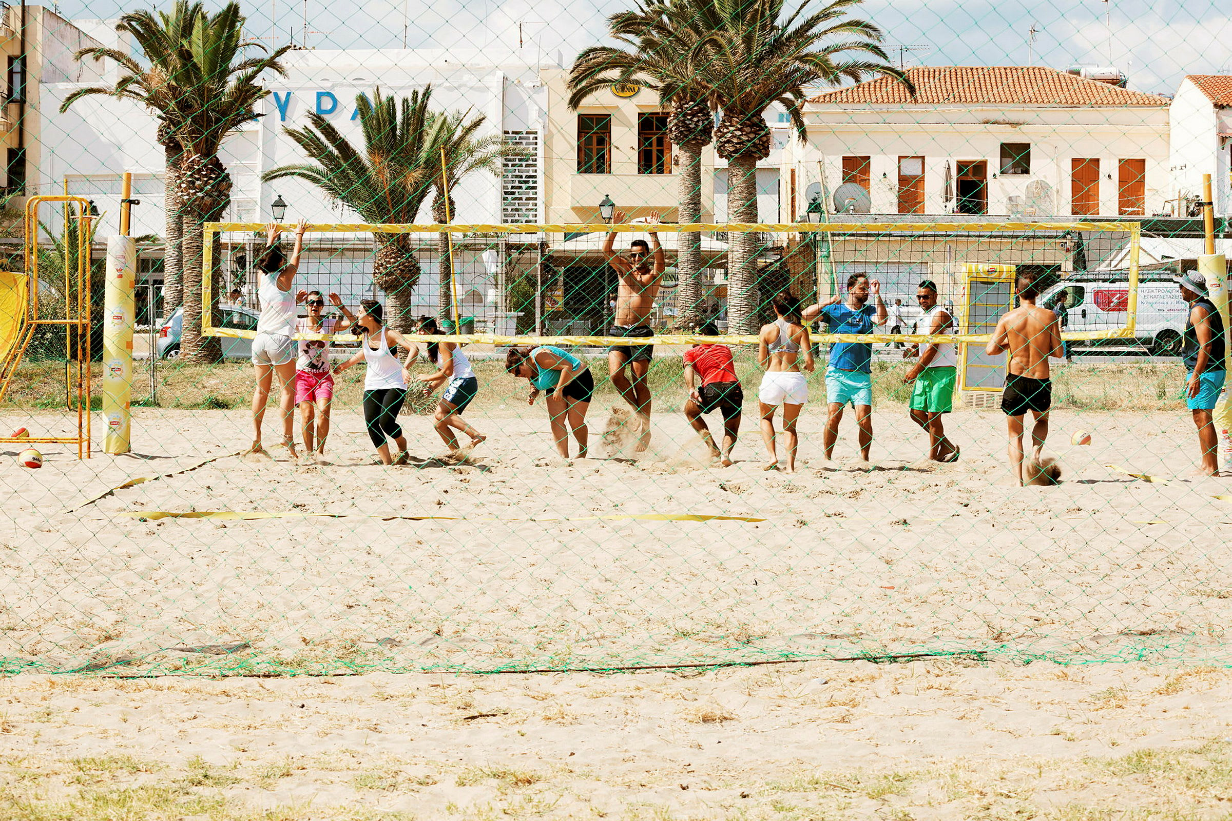 En gruppe mennesker spiller beachvolleyball på en sandbane foran bygninger og palmer. (AI-genereret tekst)