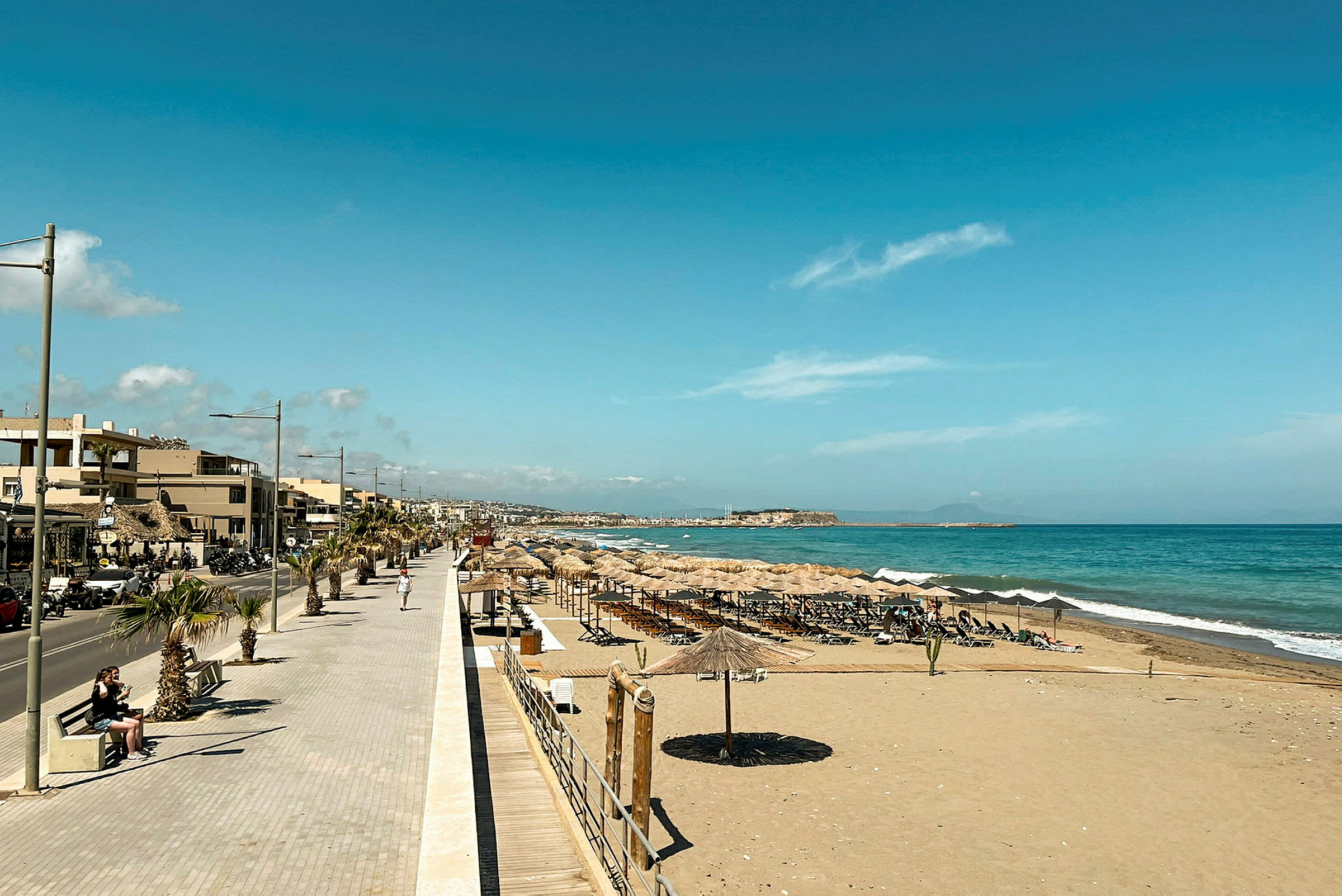 En strand med parasoller og liggestole ved siden af en strandpromenade og små bygninger. Havet ses i baggrunden. (AI-genereret tekst)