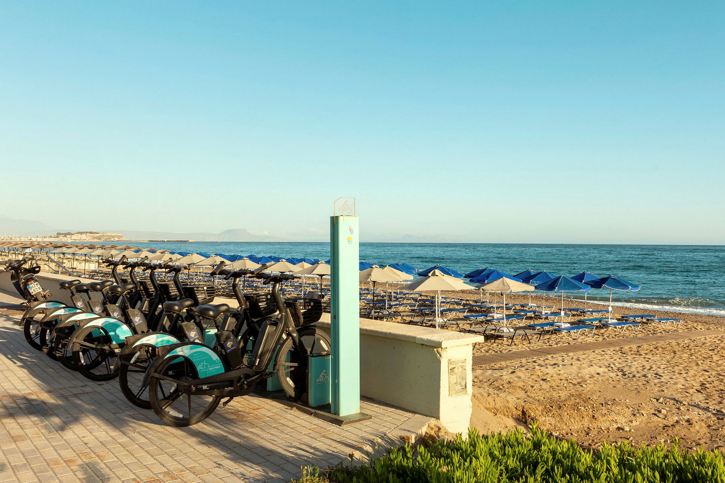 Cykler parkeret ved en strandpromenade med parasoller og liggestole ved havet. (AI-genereret tekst)