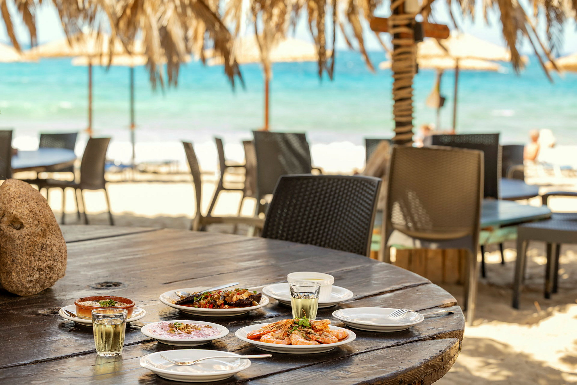 Et bord på en strandrestaurant med flere tallerkener med mad og drikke. Baggrunden viser sandstrand og havet. (AI-genereret tekst)