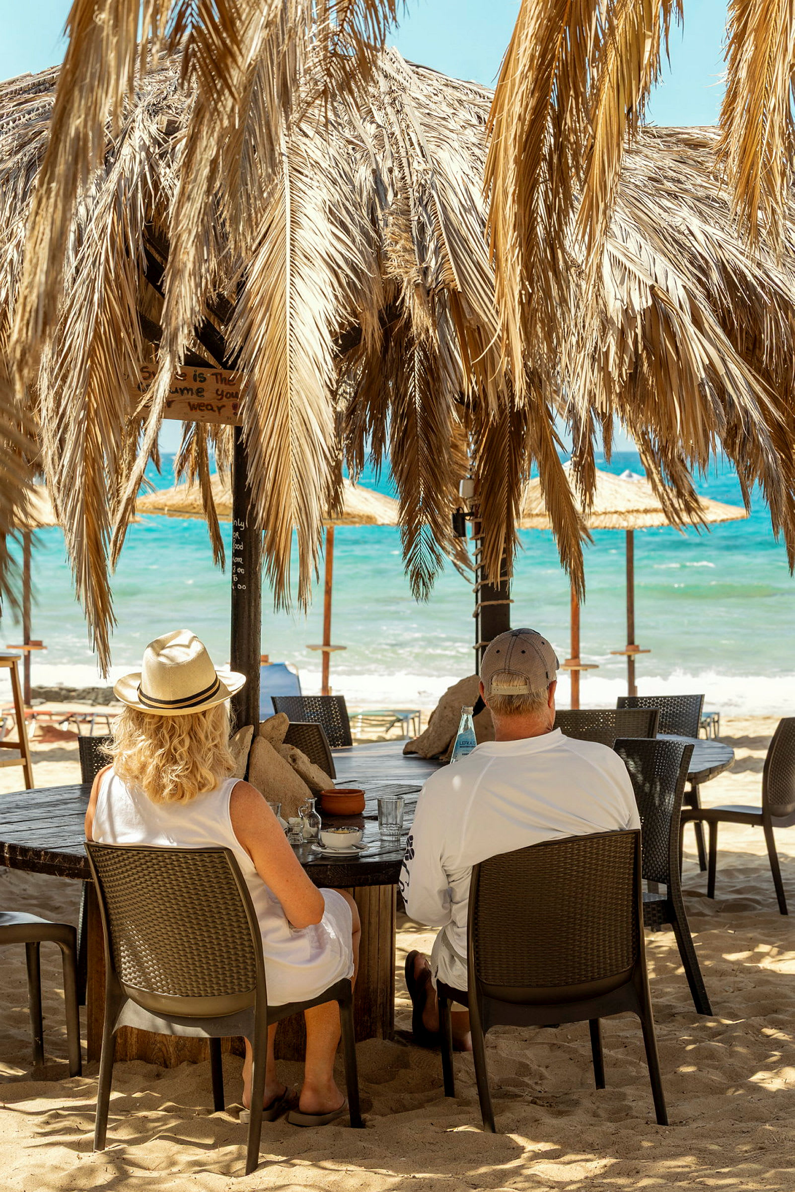 To personer sidder ved et bord under en parasol lavet af palmeblade på en strand ved havet. (AI-genereret tekst)