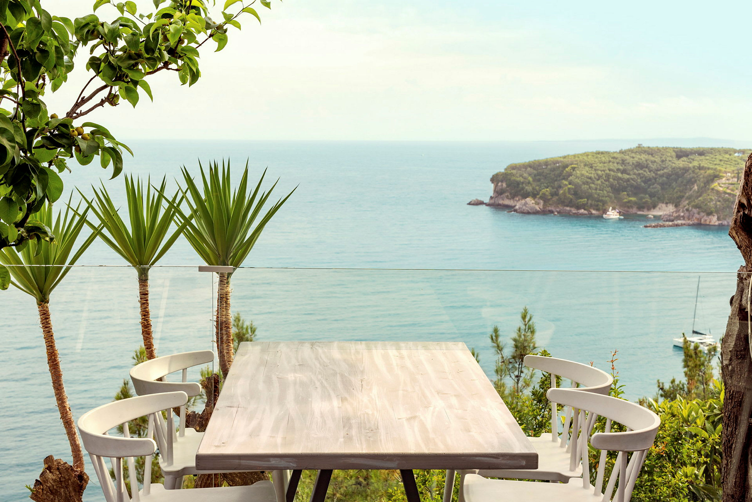 Bord med stole på en terrasse med udsigt over havet og en grøn skovklædt odde. (AI-genereret tekst)