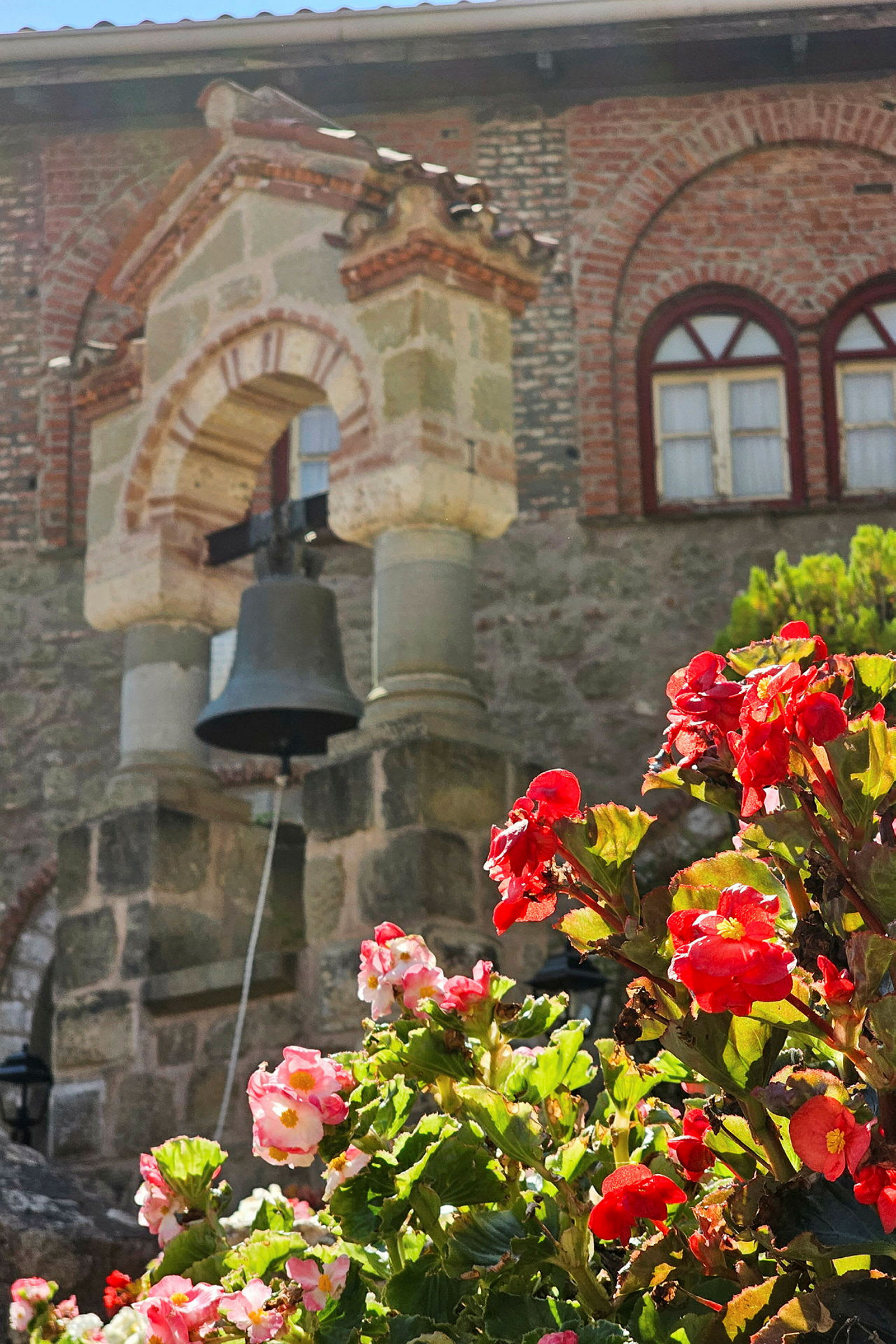 Billede af en kirkebygning med en klokke i forgrunden og røde blomster i fokus. (AI-genereret tekst)
