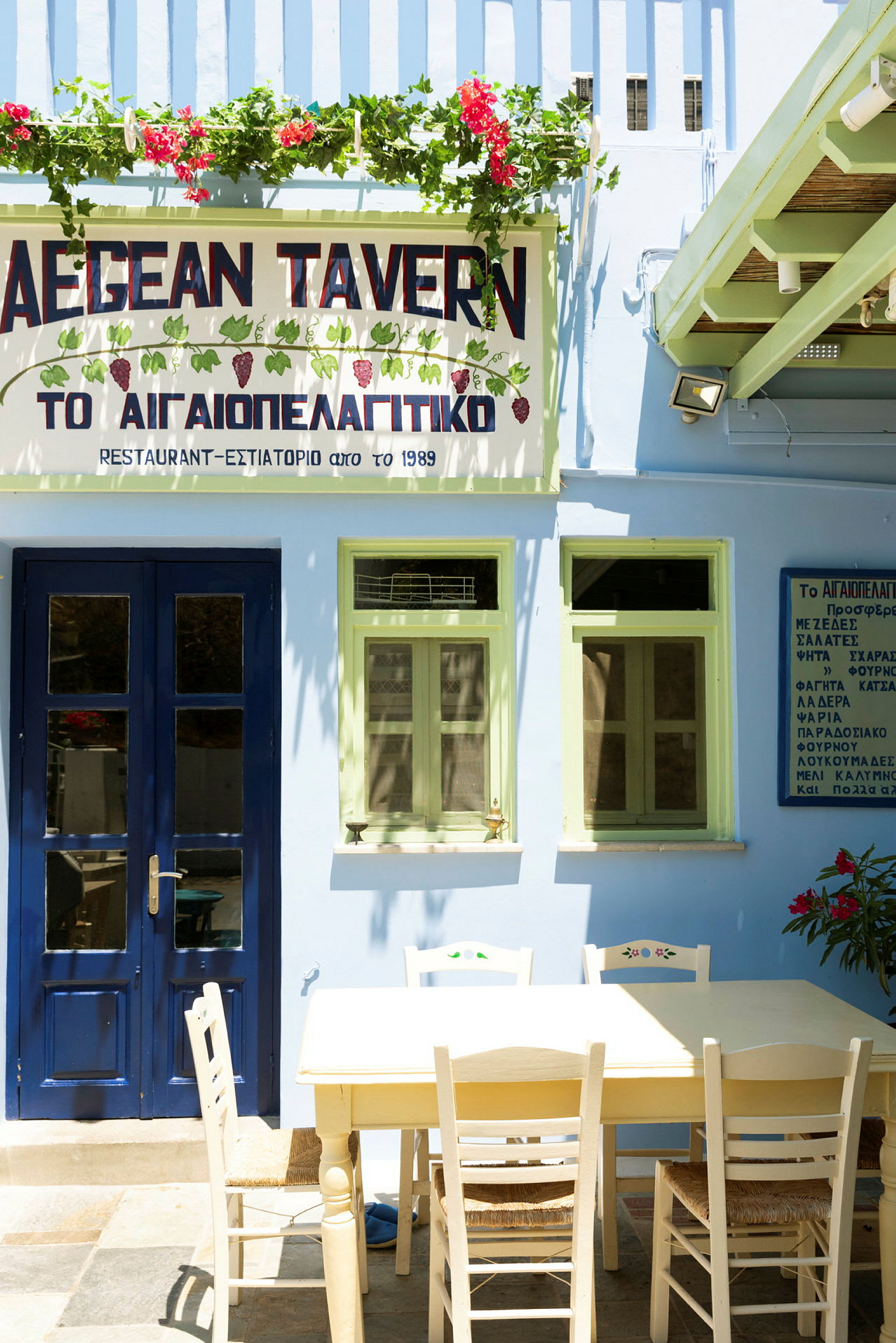 En bygning med blå dør og grønne vinduer, et skilt med teksten 'AEGEAN TAVERN' og et bord med stole foran. (AI-genereret tekst)