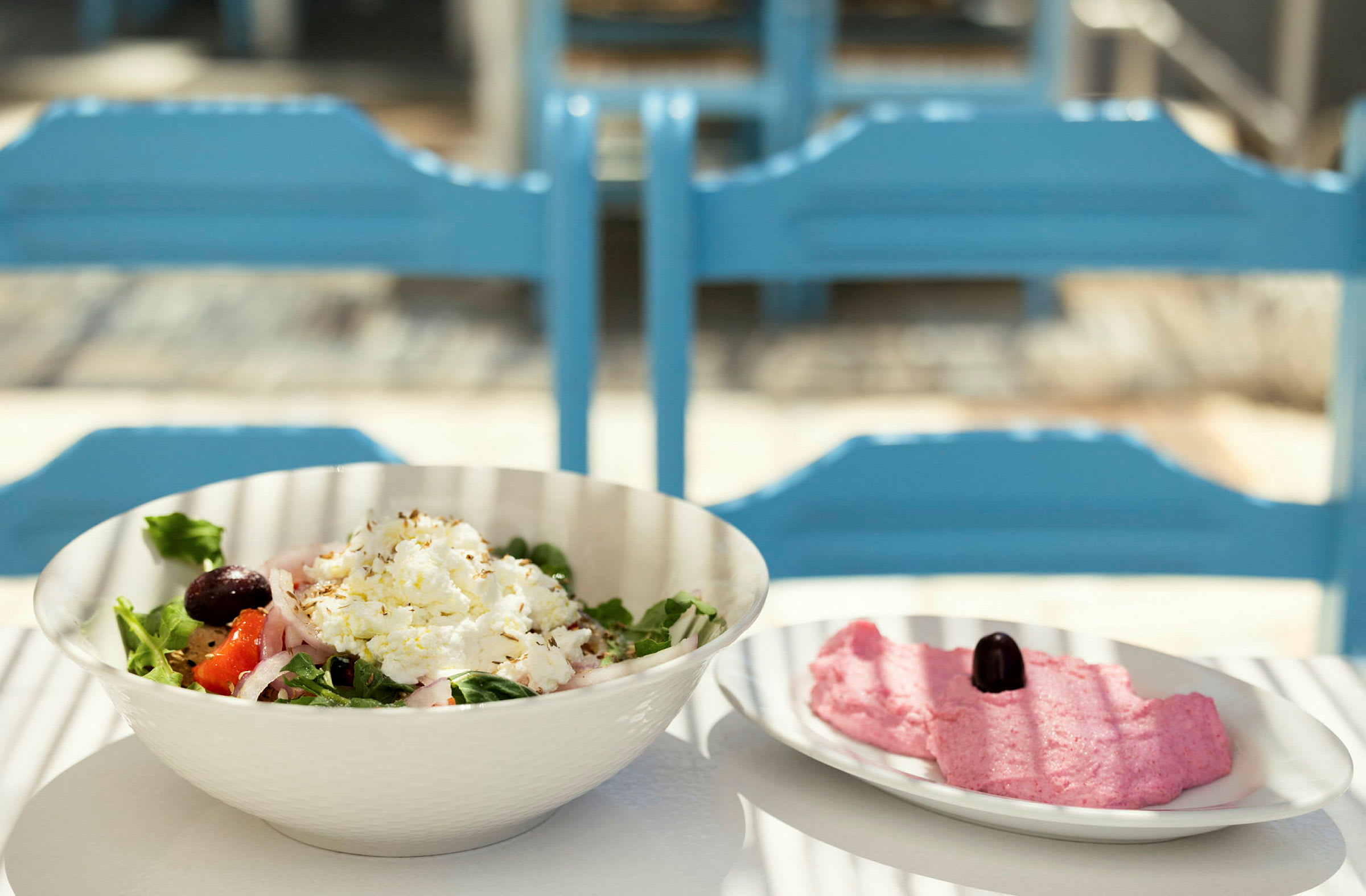 En skål med salat og en tallerken med lyserød masse samt en oliven på et bord med blå stole i baggrunden. (AI-genereret tekst)