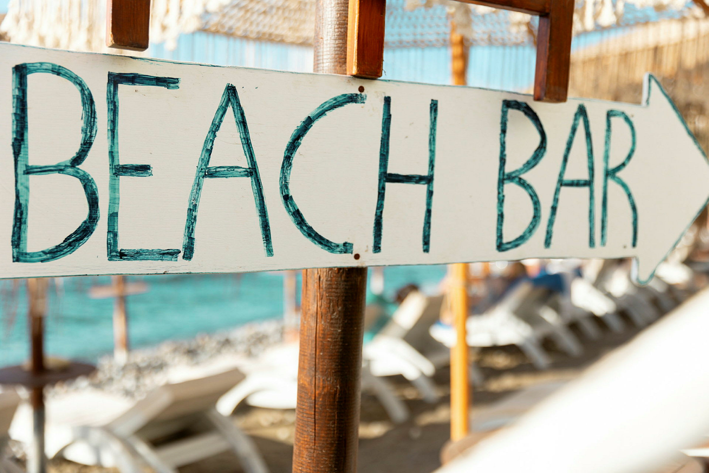Skilt med teksten 'BEACH BAR' og en pil, i baggrunden stranden med liggestole og havet. (AI-genereret tekst)