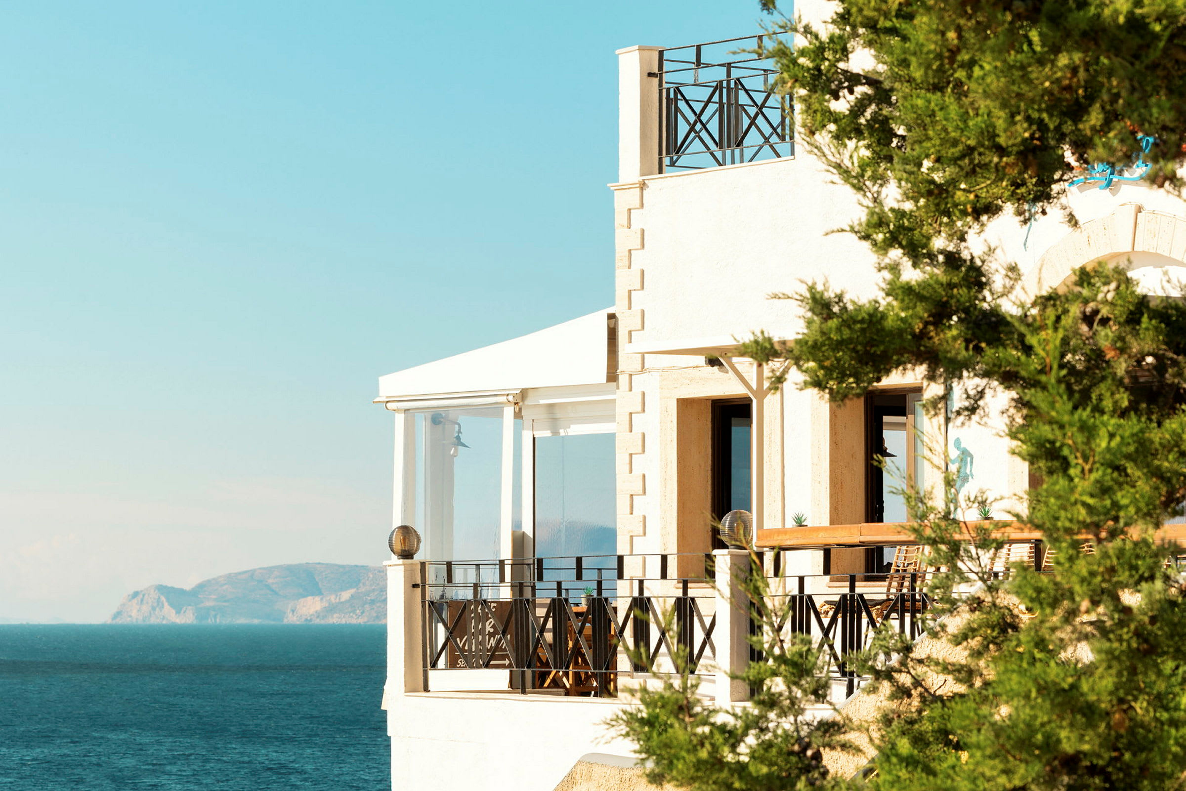 En bygning med altan ved kysten med havet og bjerge i baggrunden. (AI-genereret tekst)