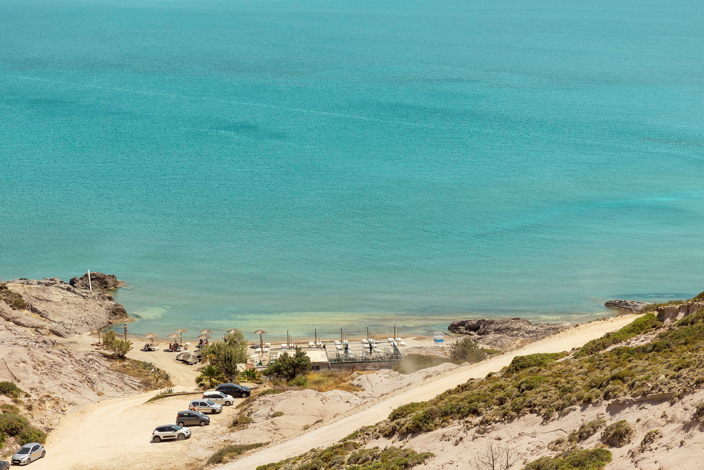 Strand med parasoller, liggestole og parkerede biler, havet i baggrunden. (AI-genereret tekst)