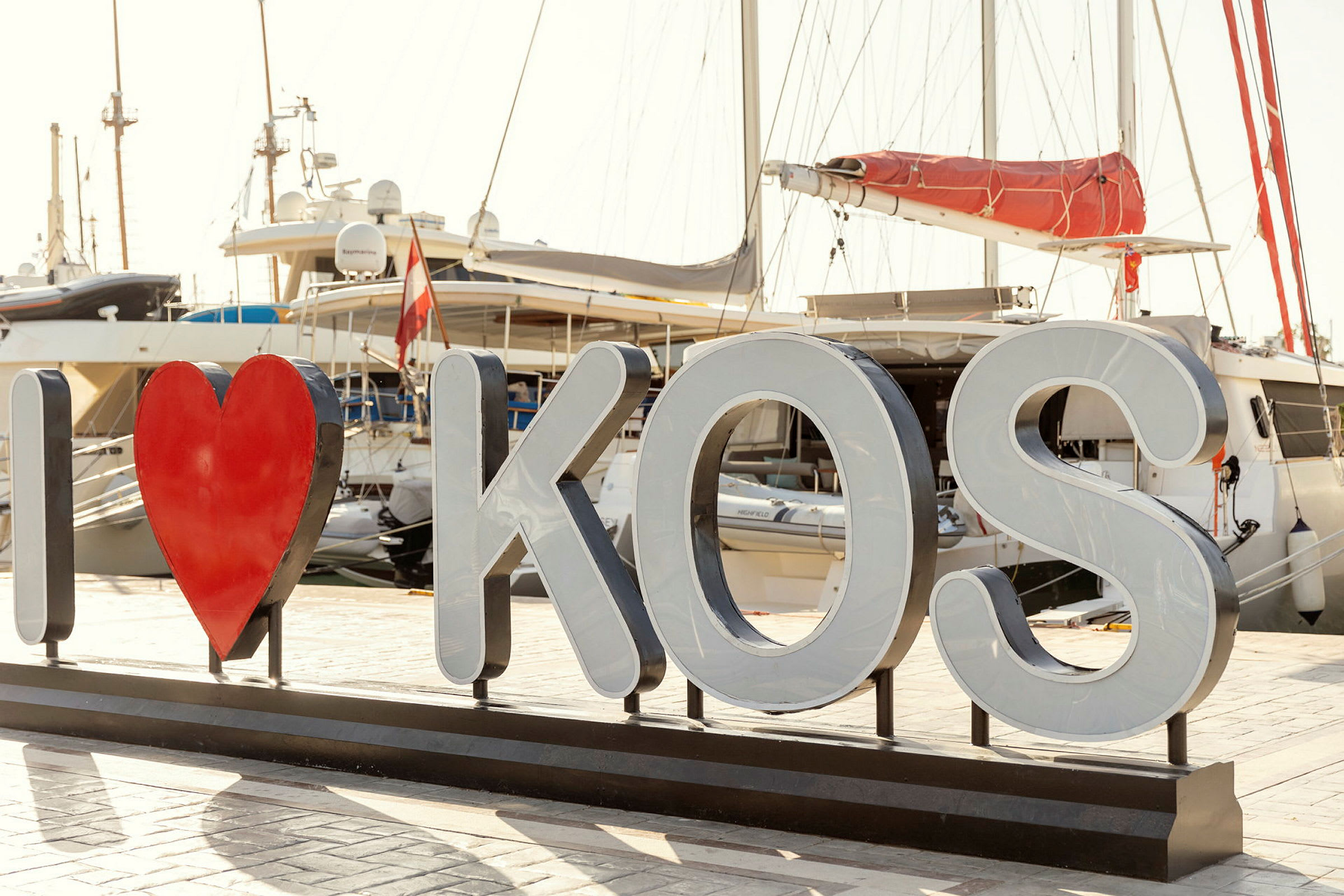 Skilt med teksten 'I ♥ KOS' foran både i en havn. (AI-genereret tekst)