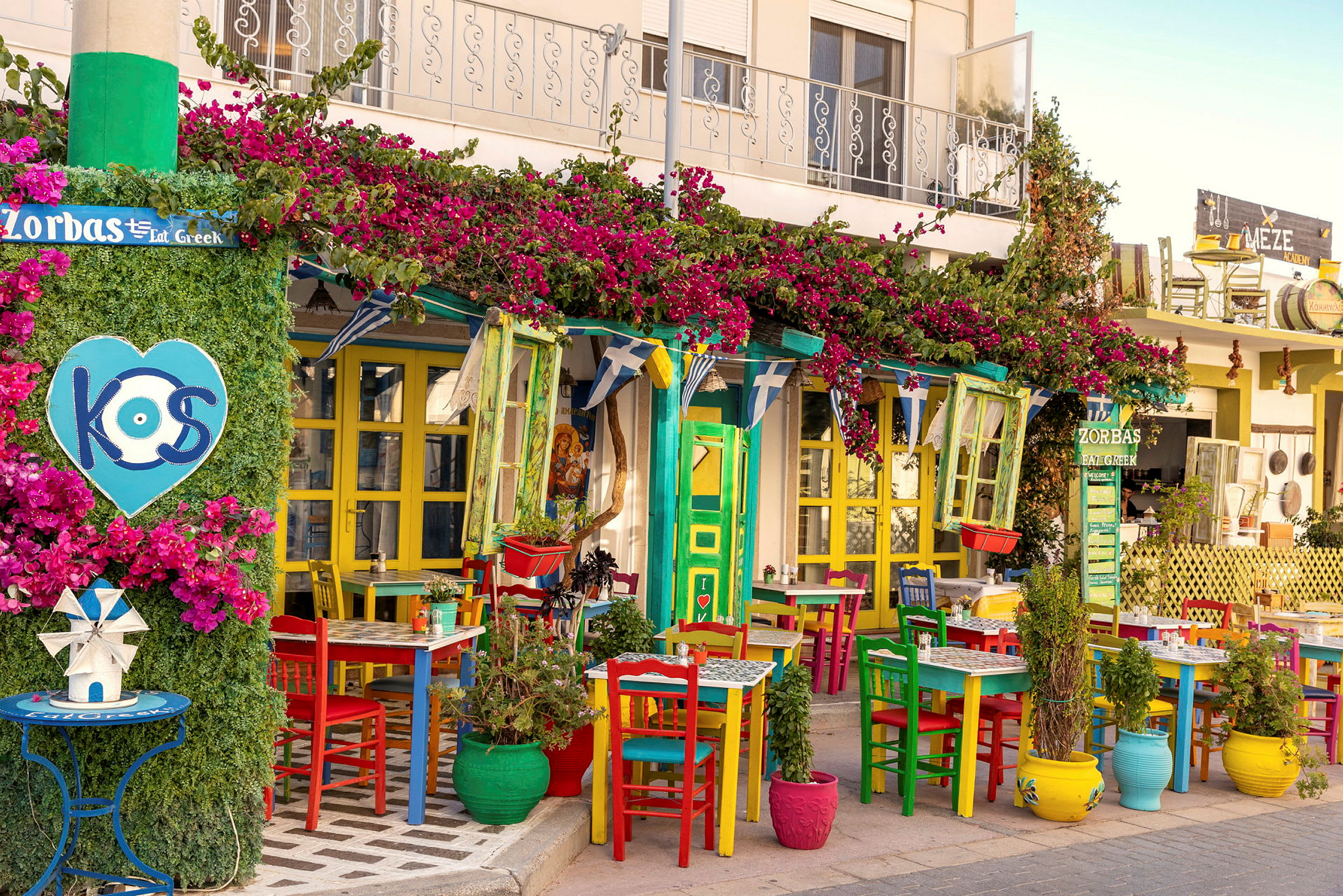 Farverige stole og borde foran en restaurant dekoreret med blomster og planter. (AI-genereret tekst)