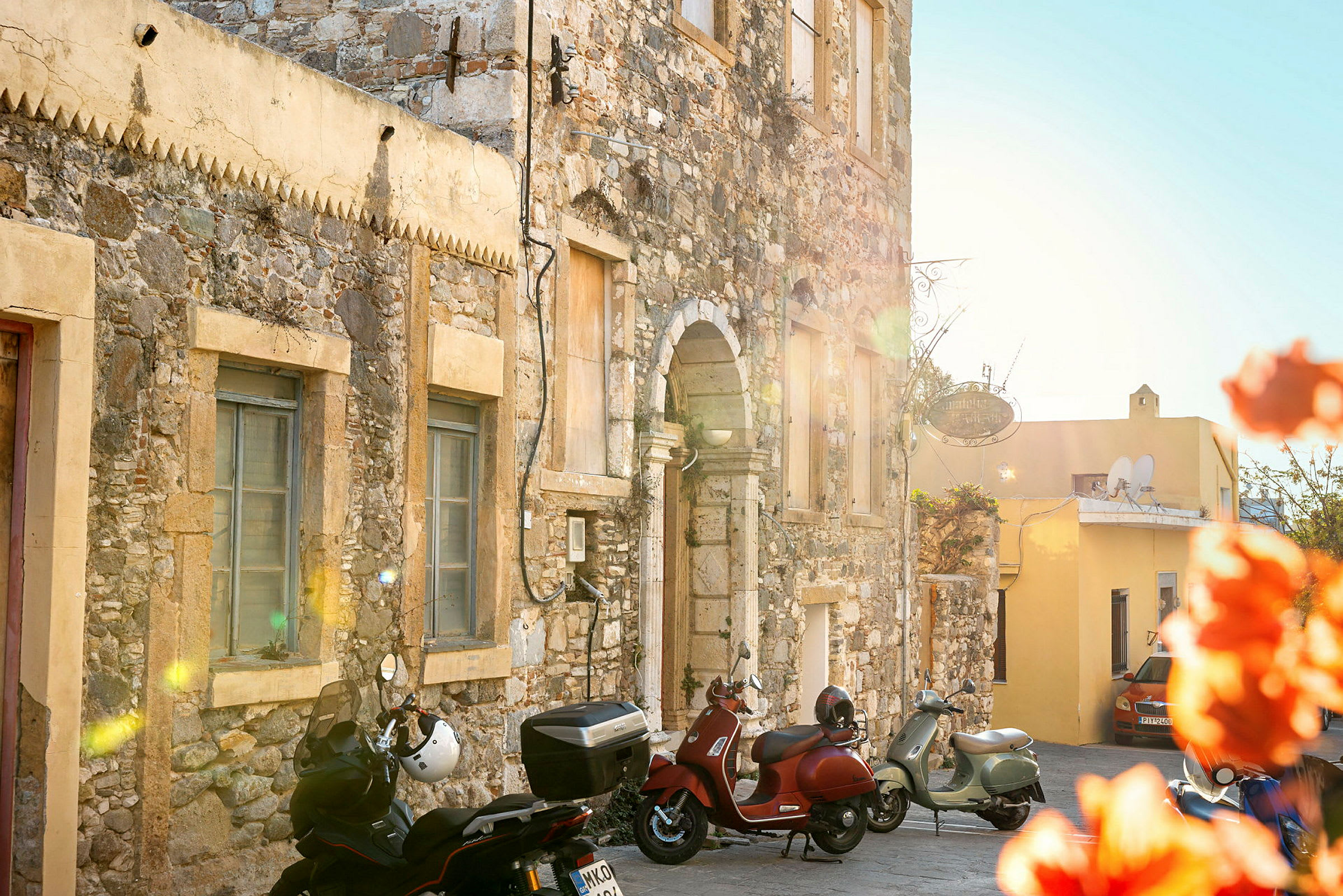 En stenbygning med vinduer og en bue, motorcykler parkeret udenfor, og en solbelyst gul bygning i baggrunden. (AI-genereret tekst)