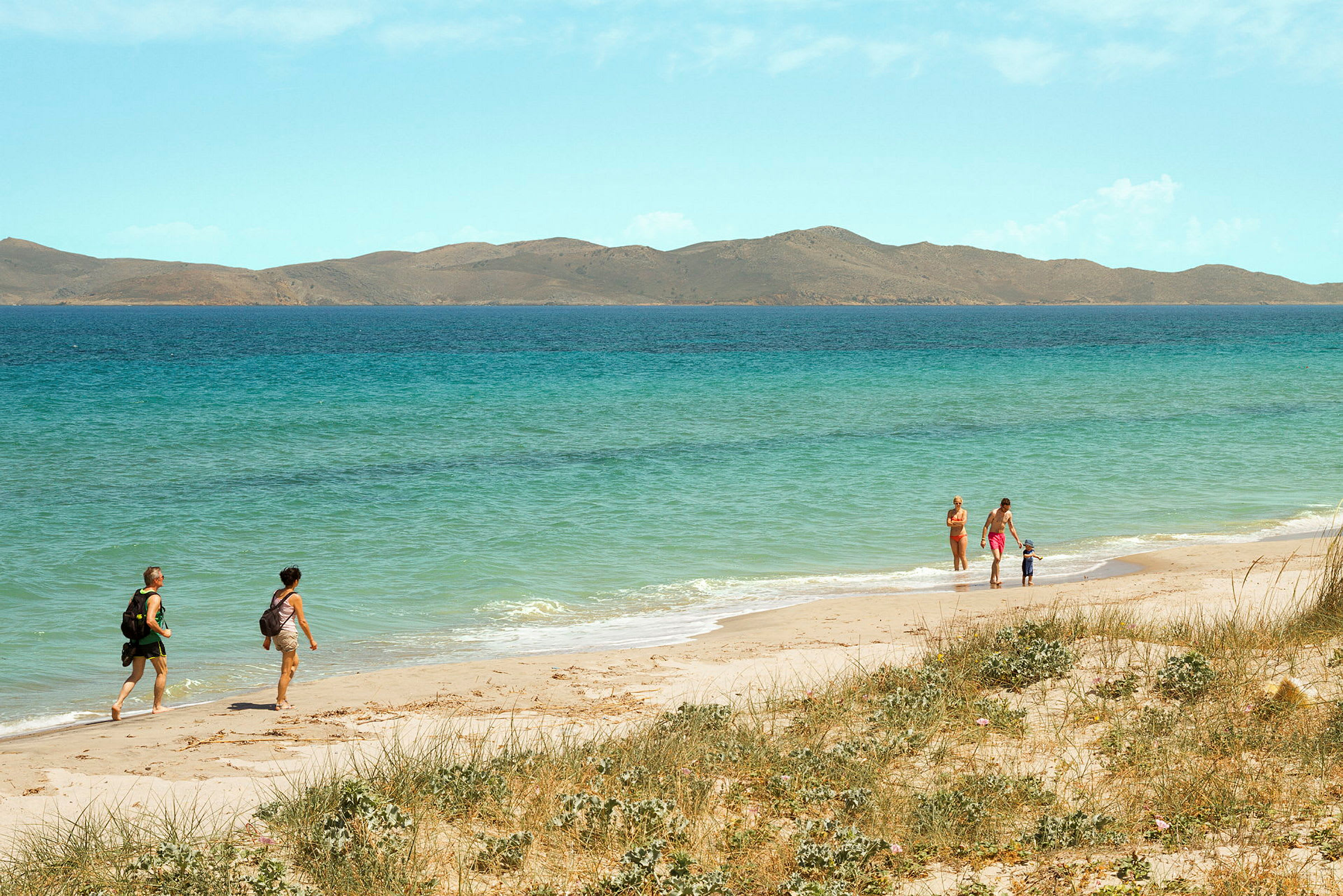 Personer går på en solrig strand med klart blåt hav og bjerge i baggrunden. (AI-genereret tekst)