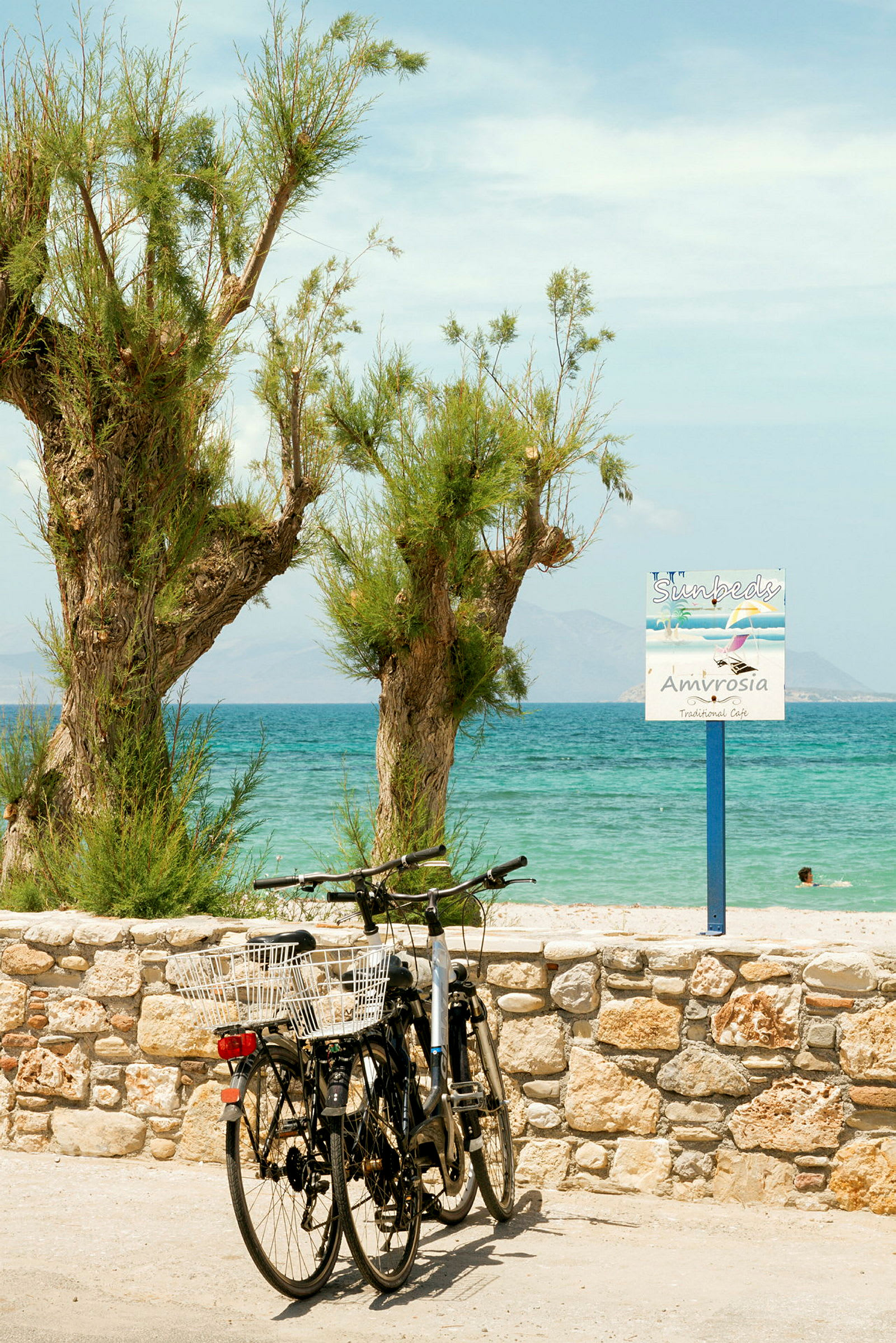 To cykler står parkeret ved et stenhegn foran træer og havet. (AI-genereret tekst)