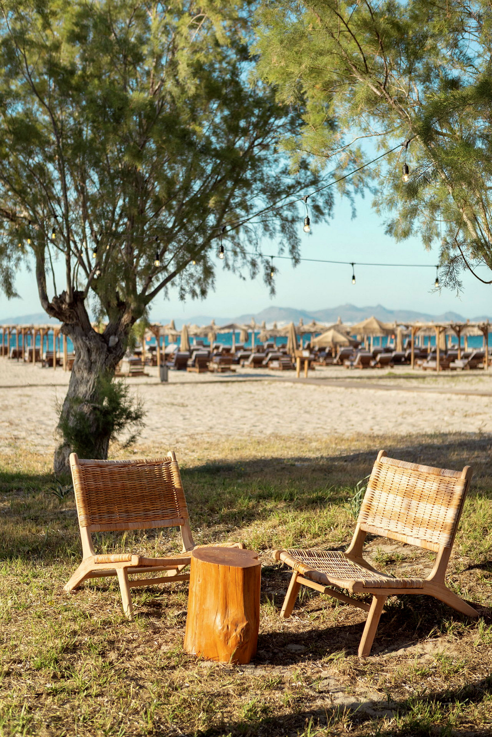 To fletstole og et lille træbord står på græs nær en strand med parasoller. (AI-genereret tekst)