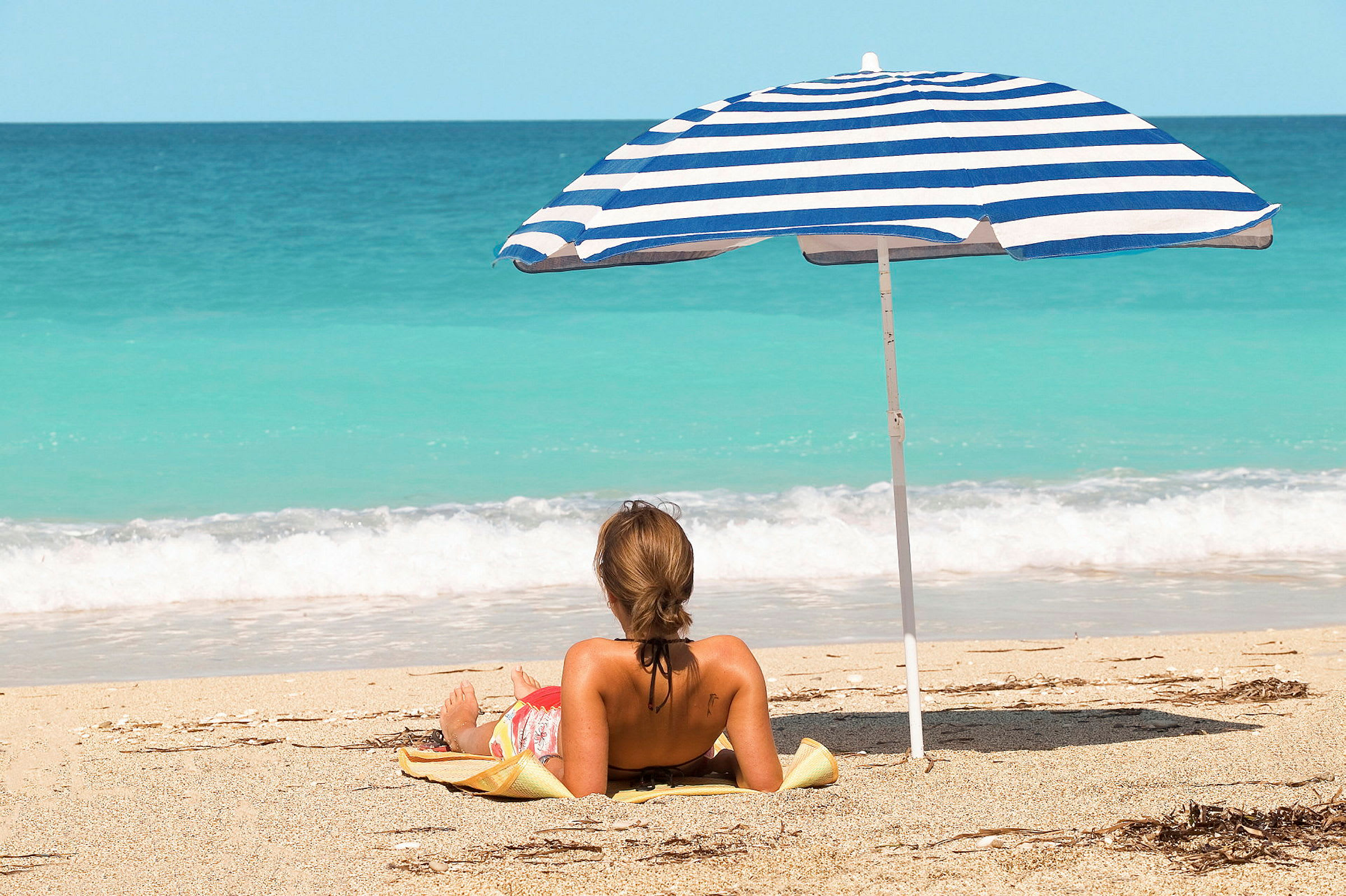 En person ligger på en strand under en blå og hvid parasol tæt på havet. (AI-genereret tekst)
