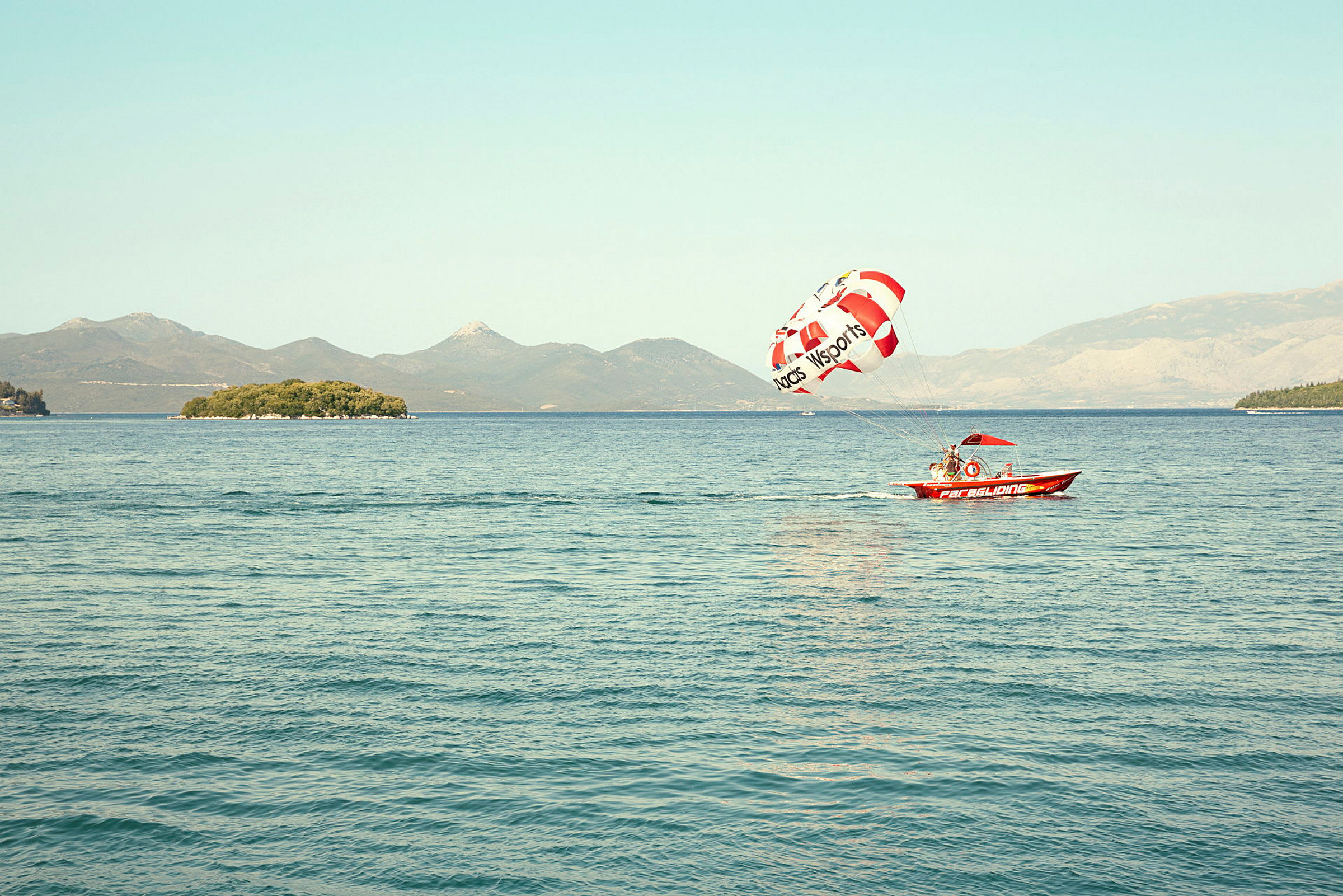 En lille motorbåd trækker en parasailing skærm over vandet med en ø i baggrunden. (AI-genereret tekst)