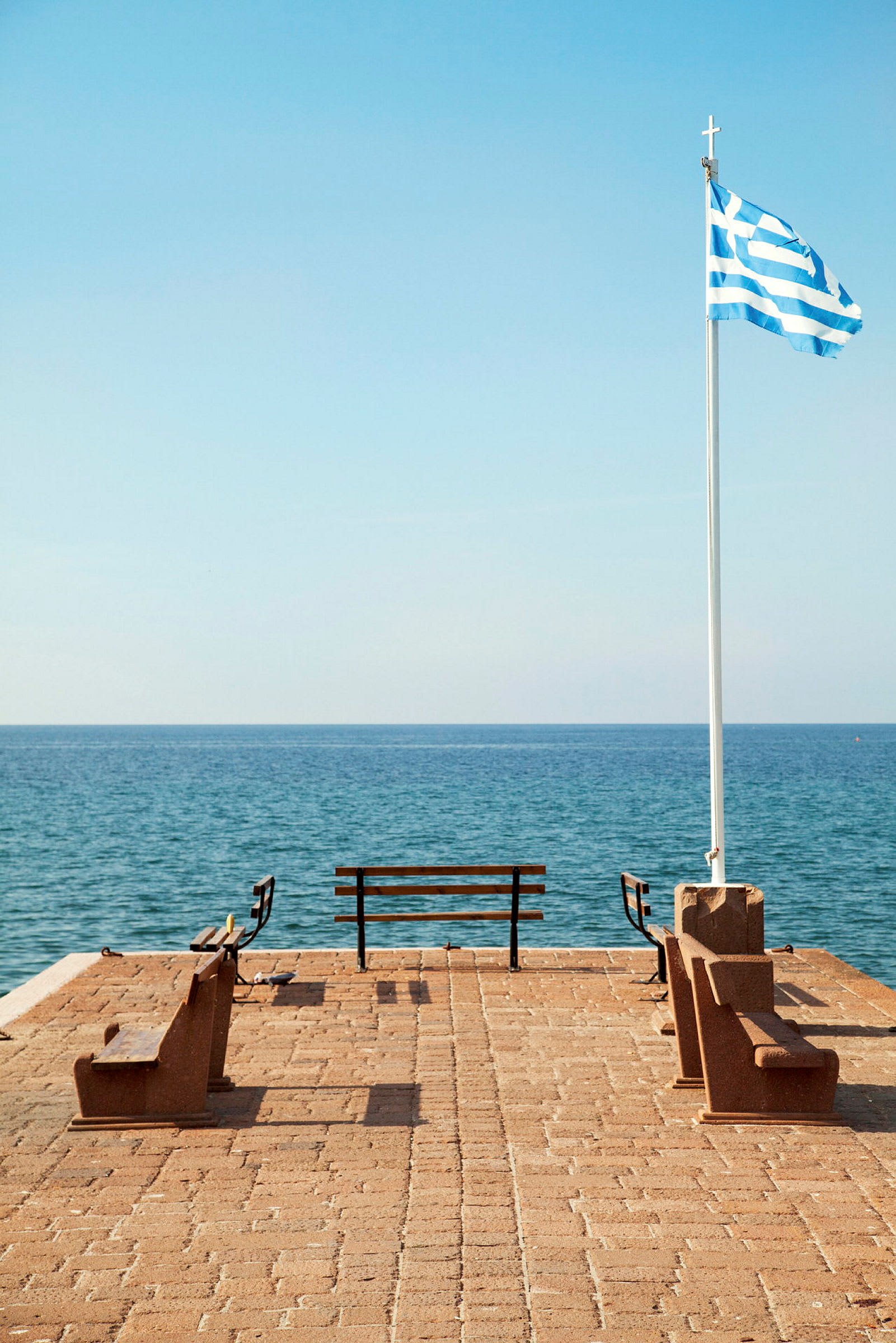 Bro med bænke og græsk flag ved havet. (AI-genereret tekst)