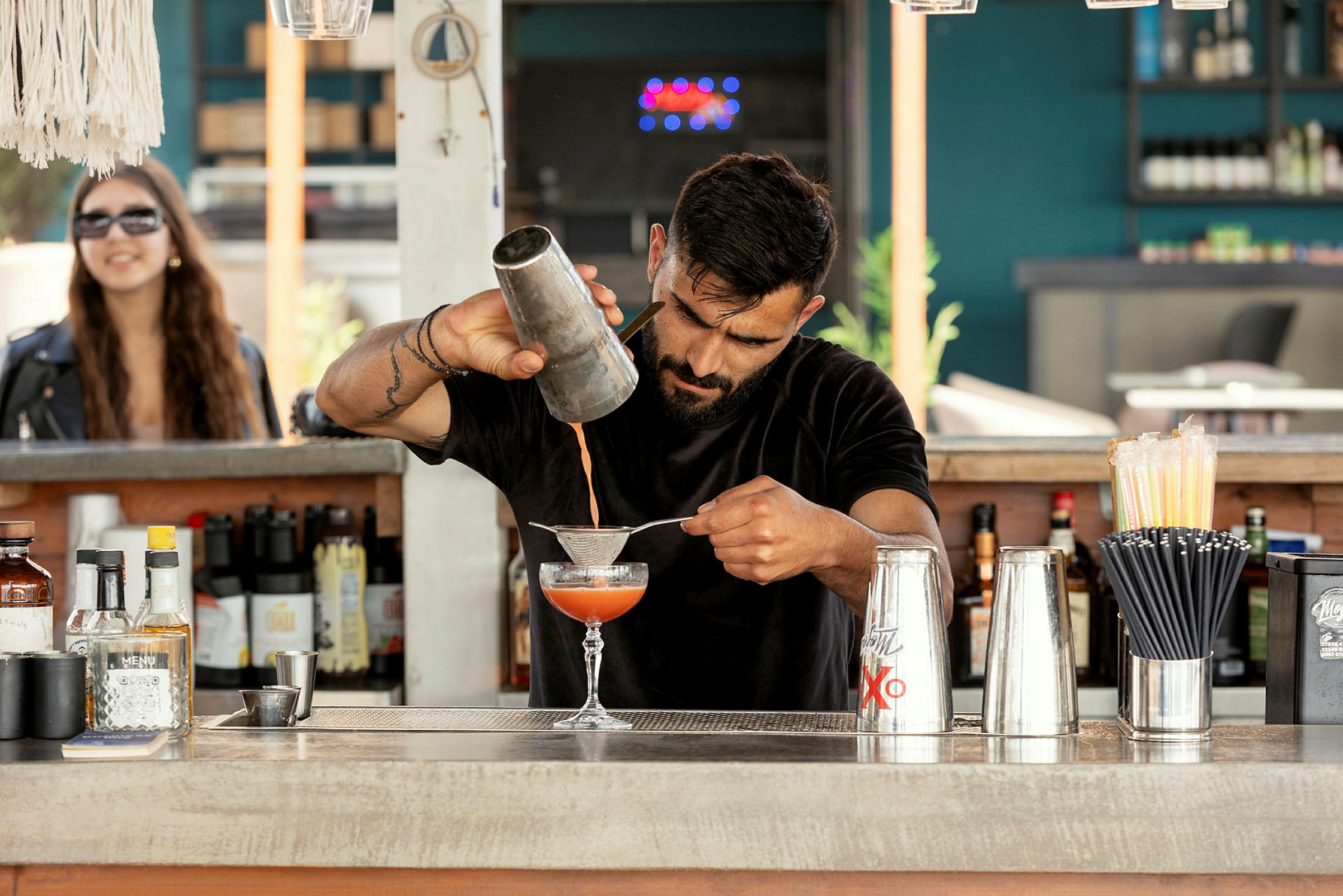 En bartender ryster og silar en cocktail ved siden af flasker, stålrystere og sugerør på en udendørs bar. (AI-genereret tekst)