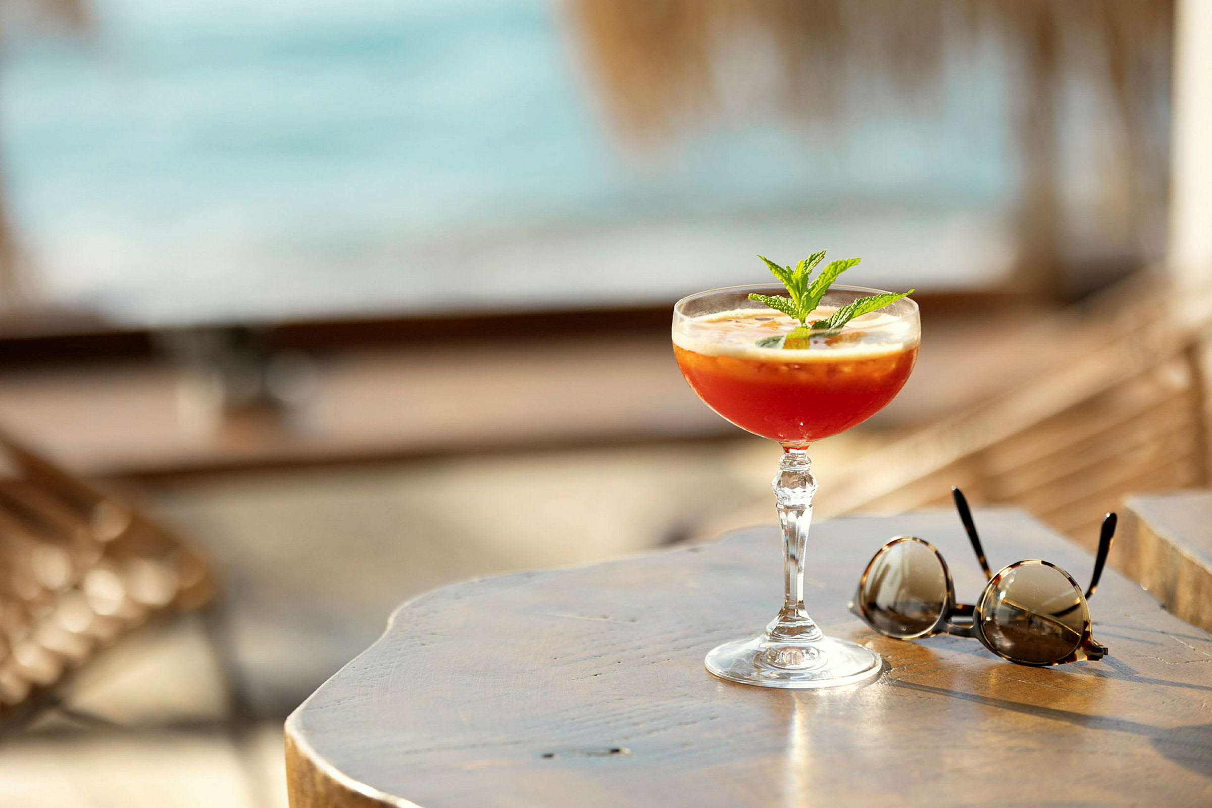 En cocktail og et par solbriller på et træbord ved stranden. (AI-genereret tekst)