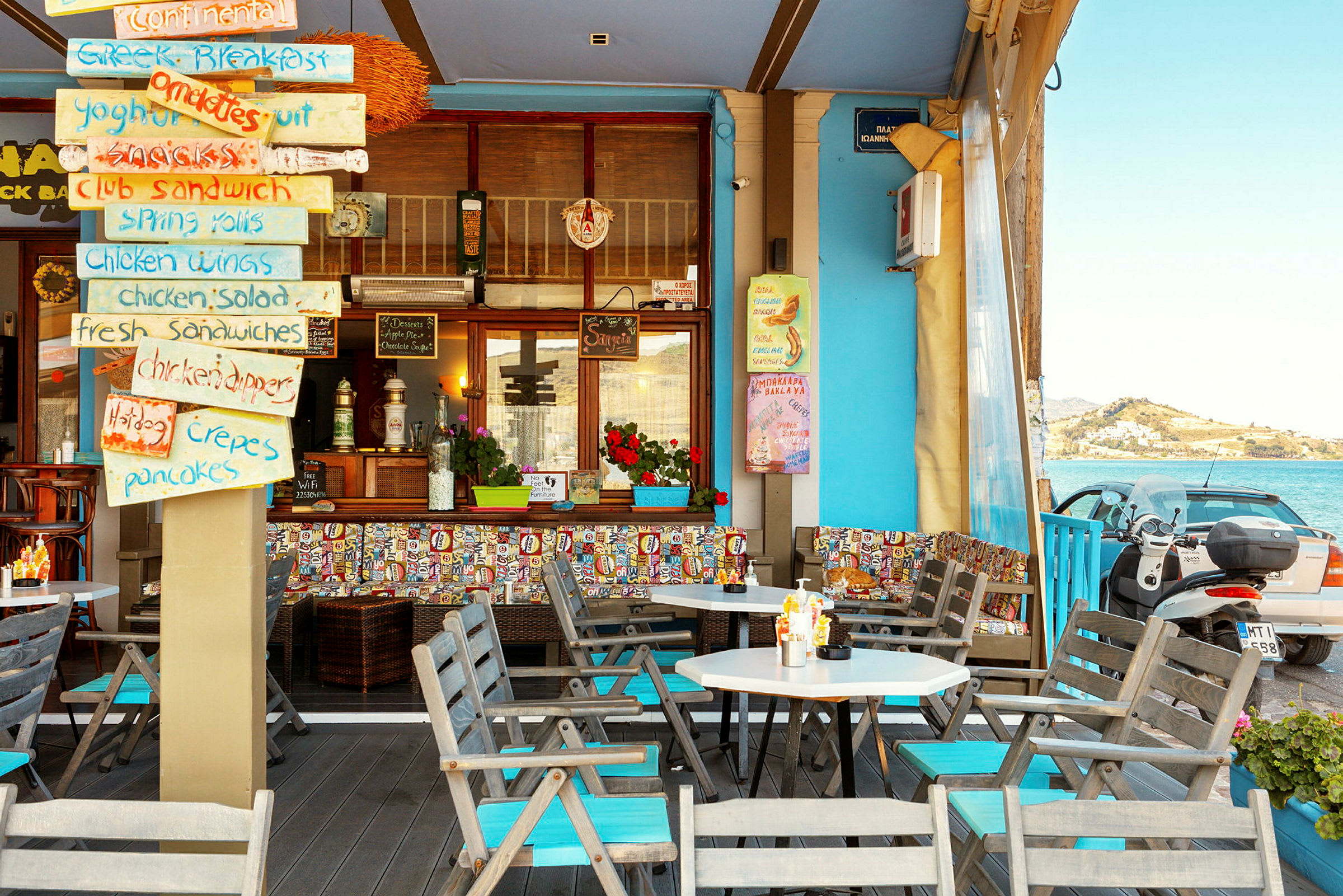 En farverig caféterrasse med skilte der viser menuen, siddepladser og udsigt over havet. (AI-genereret tekst)