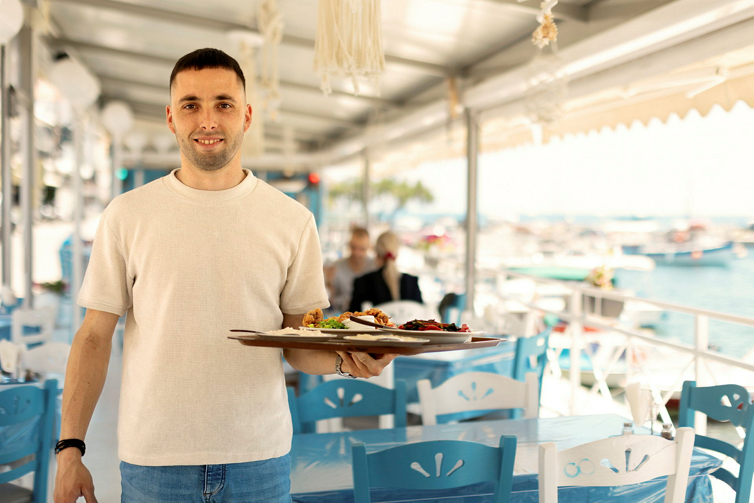 Personen holder en tallerken med mad på en udendørs restaurant ved havet. (AI-genereret tekst)