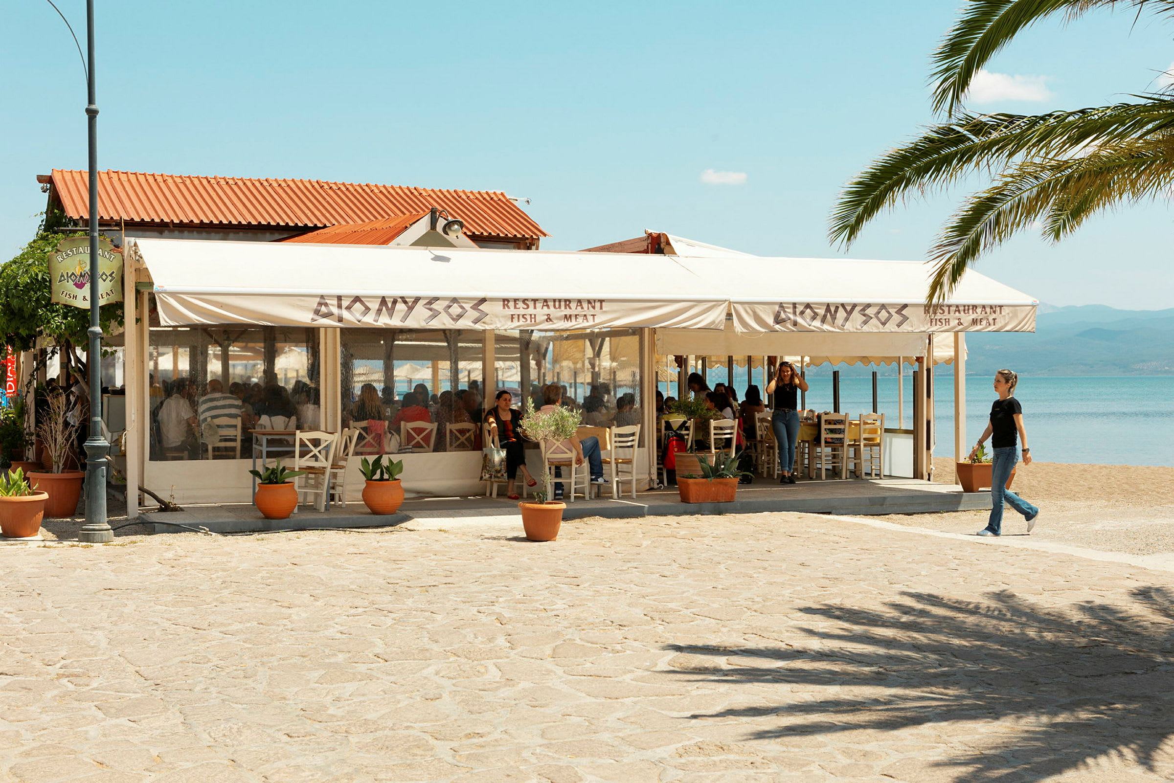 En restaurant ved stranden med skilte, der angiver, at den serverer fisk og kød. Udeserveringen er dækket med parasoller. (AI-genereret tekst)