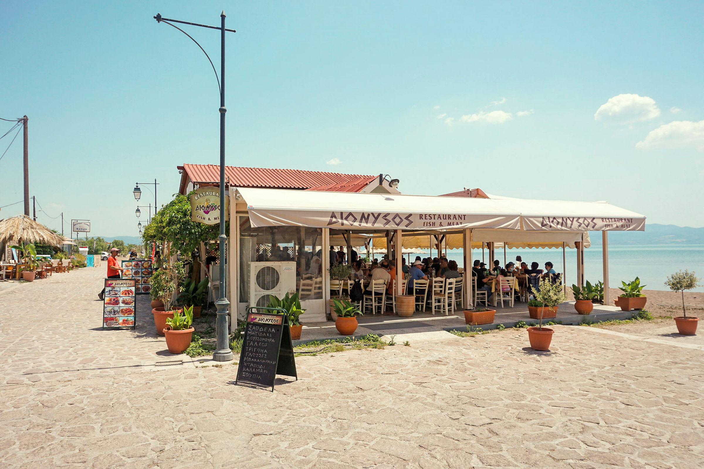 En udendørsrestaurant med siddepladser under et stort telt. I baggrunden ses havet og en klar himmel. (AI-genereret tekst)