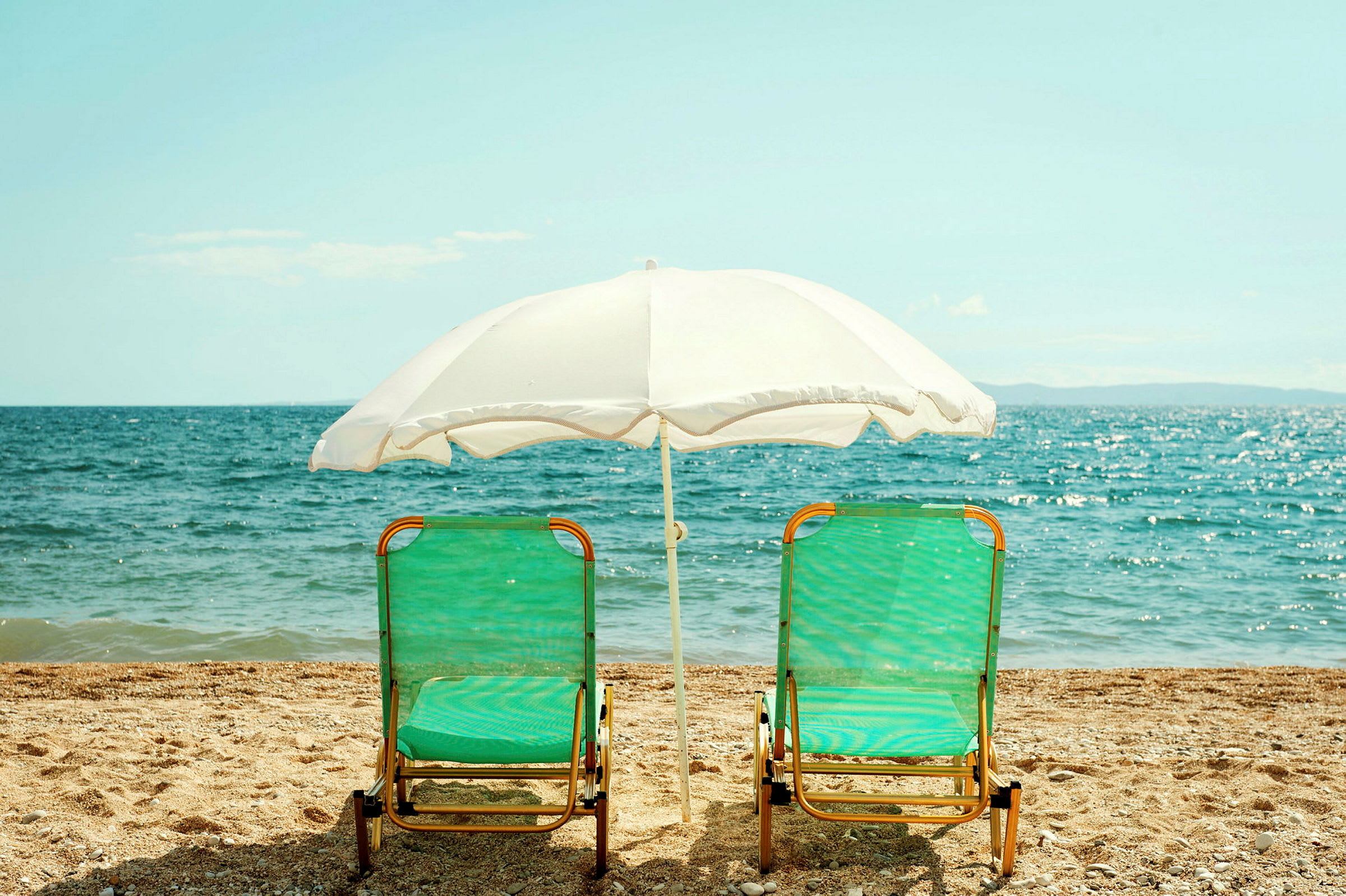 To liggestole med grønne sæder og en hvid parasol står på stranden foran havet. (AI-genereret tekst)