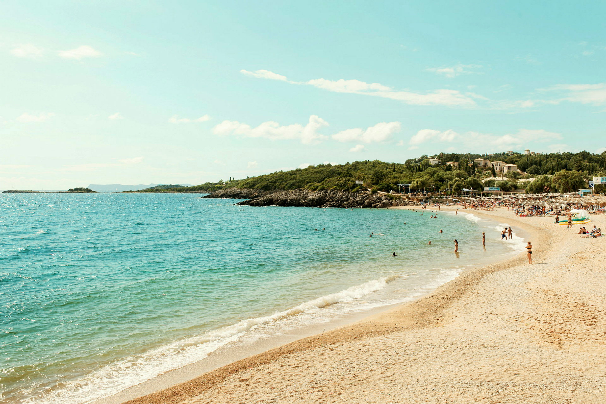 Strand med sand og klart blåt vand, omgivet af grønt og folk der bader. (AI-genereret tekst)