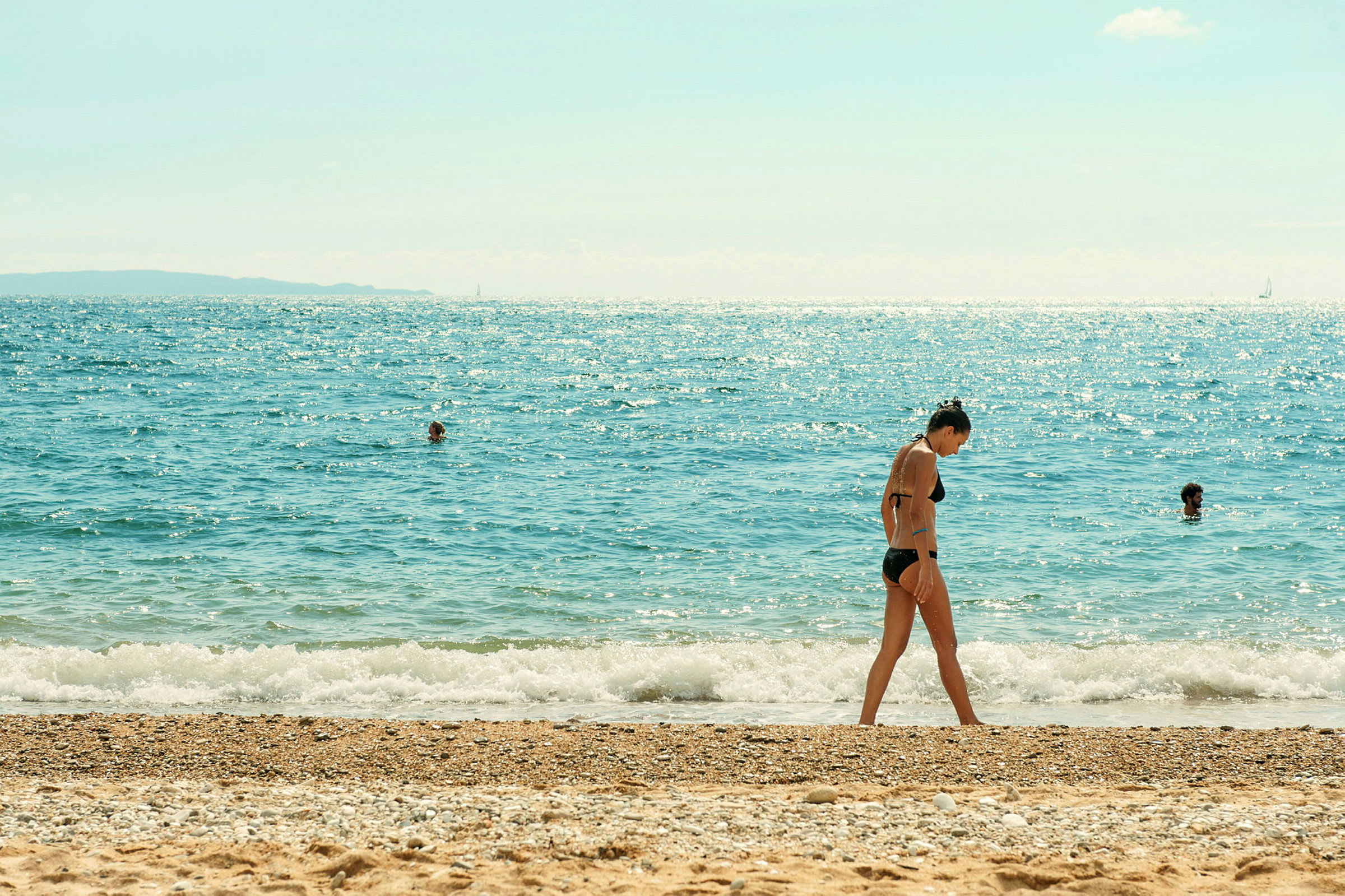 En strand med sand og sten, og havet i baggrunden med personer, der bader. (AI-genereret tekst)