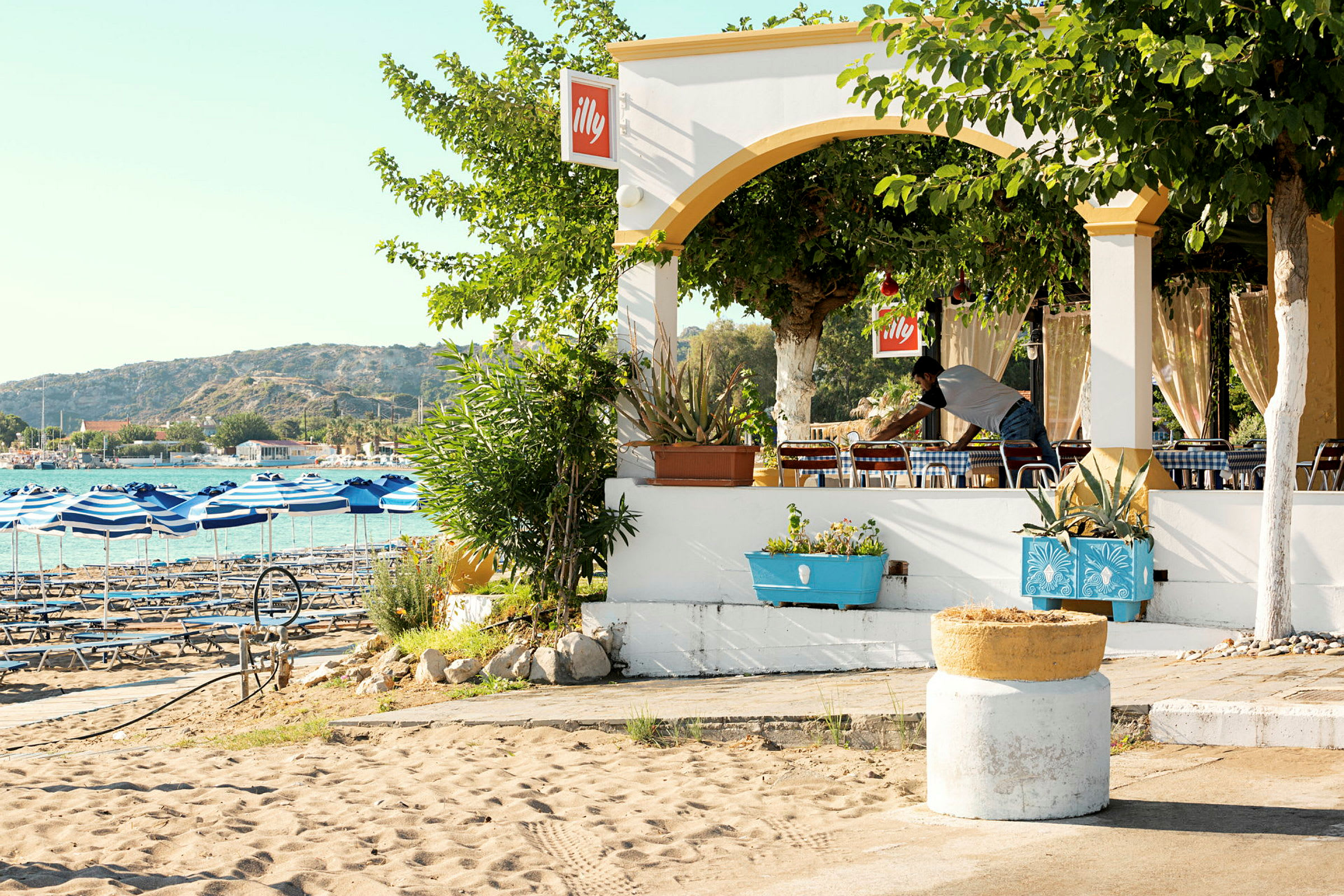 En strandnær restaurant med blå- og hvidstribede parasoller ved havet og omgivelser med grønt. (AI-genereret tekst)