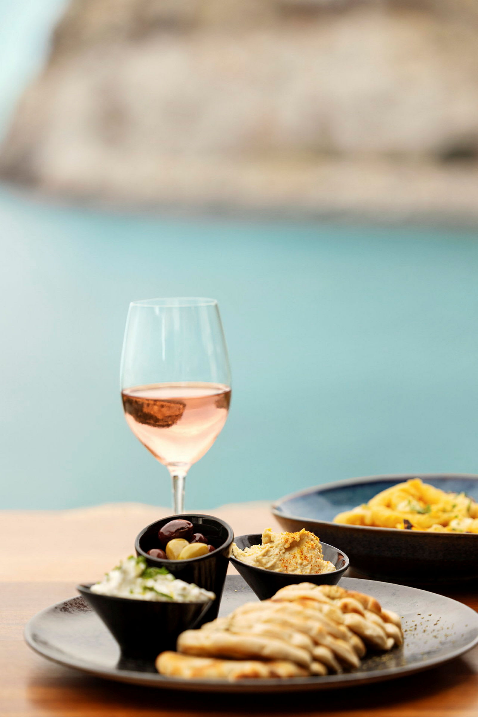 Et glas rosévin ved siden af en tallerken med brødstykker, en skål med oliven og cremede dips. (AI-genereret tekst)
