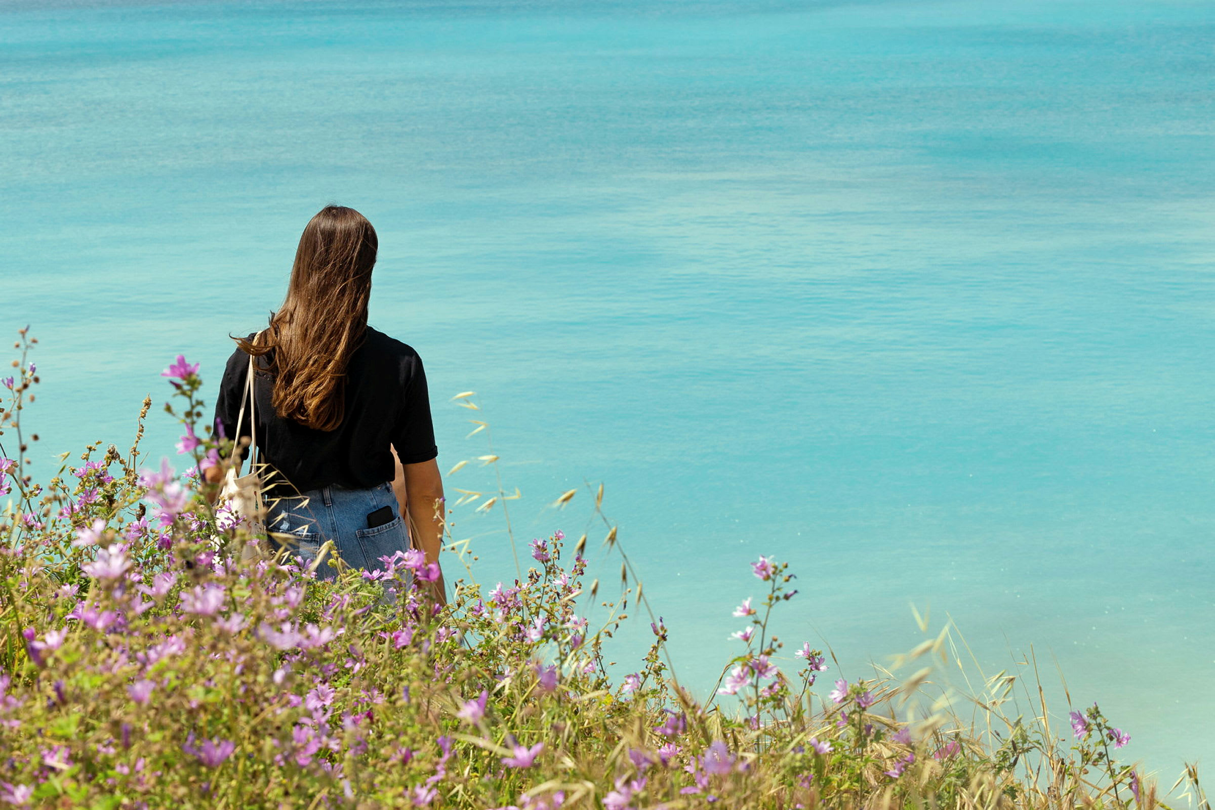 En kvinde står ved kysten med ryggen vendt mod kameraet og kigger ud over havet, omgivet af blomstrende planter. (AI-genereret tekst)