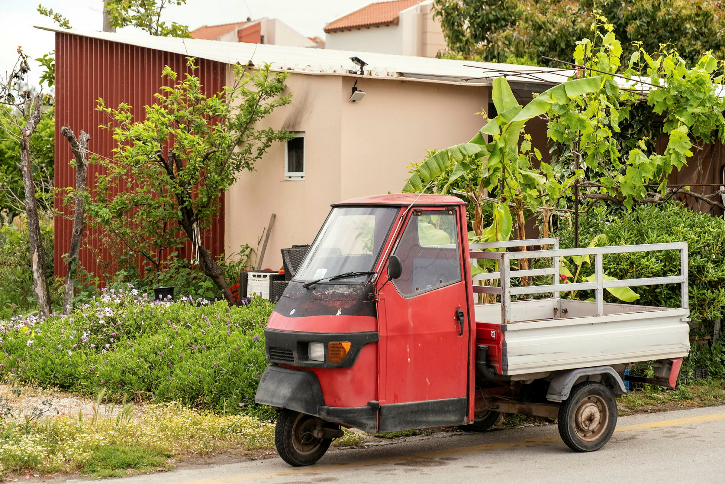 En rød trehjulet lastbil er parkeret foran et hus med træer og planter omkring. (AI-genereret tekst)