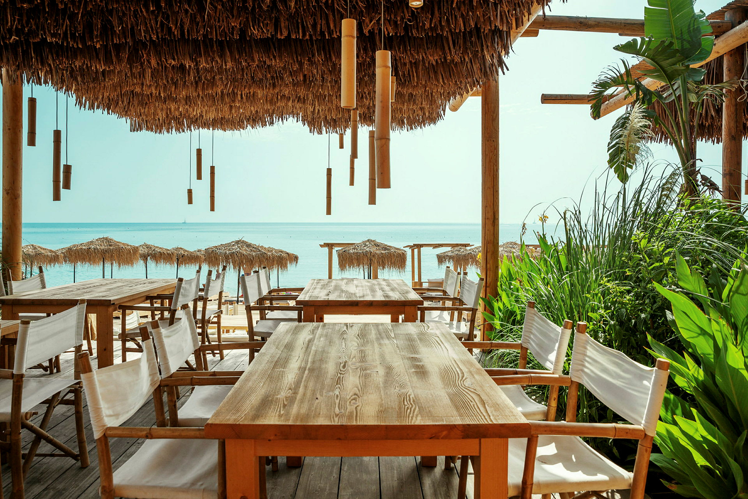 Udendørs serveringsområde med træborde og stole under en pergola med stråtag, med udsigt over stranden og havet. (AI-genereret tekst)