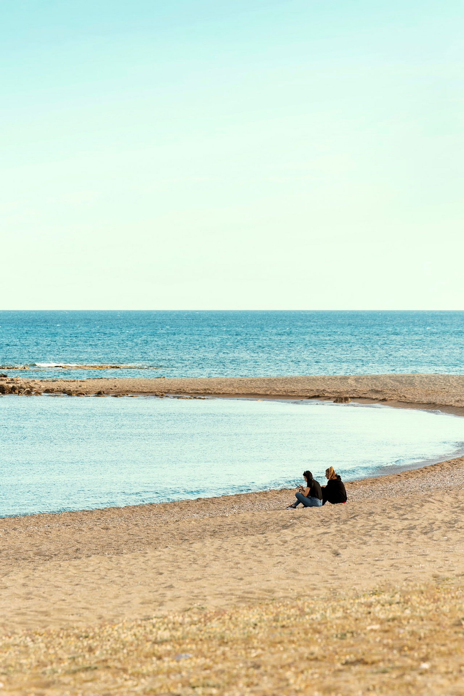To personer sidder på en strand tæt på havet under klar himmel. (AI-genereret tekst)